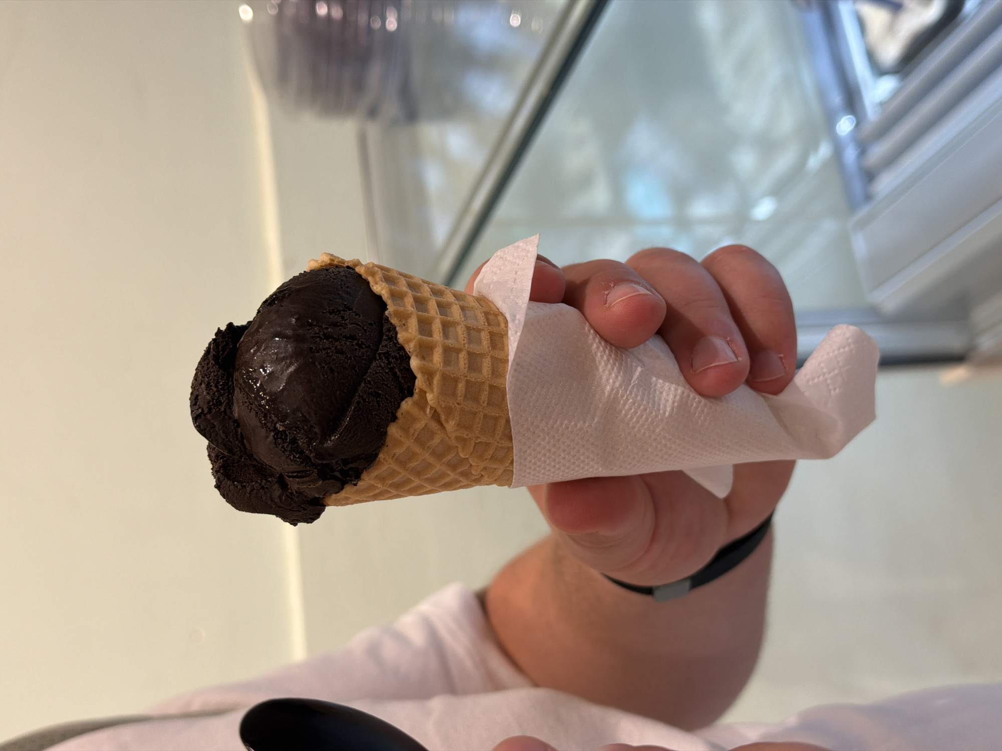 A hand holds a chocolate ice cream cone wrapped in a white napkin. The waffle cone is light brown, and the ice cream appears to be dark chocolate. A black bracelet is visible on the wrist of the person holding the cone. The background shows a reflective surface, possibly glass or a shiny countertop. A white wall is also visible. The image was taken in Hydra, Greece.