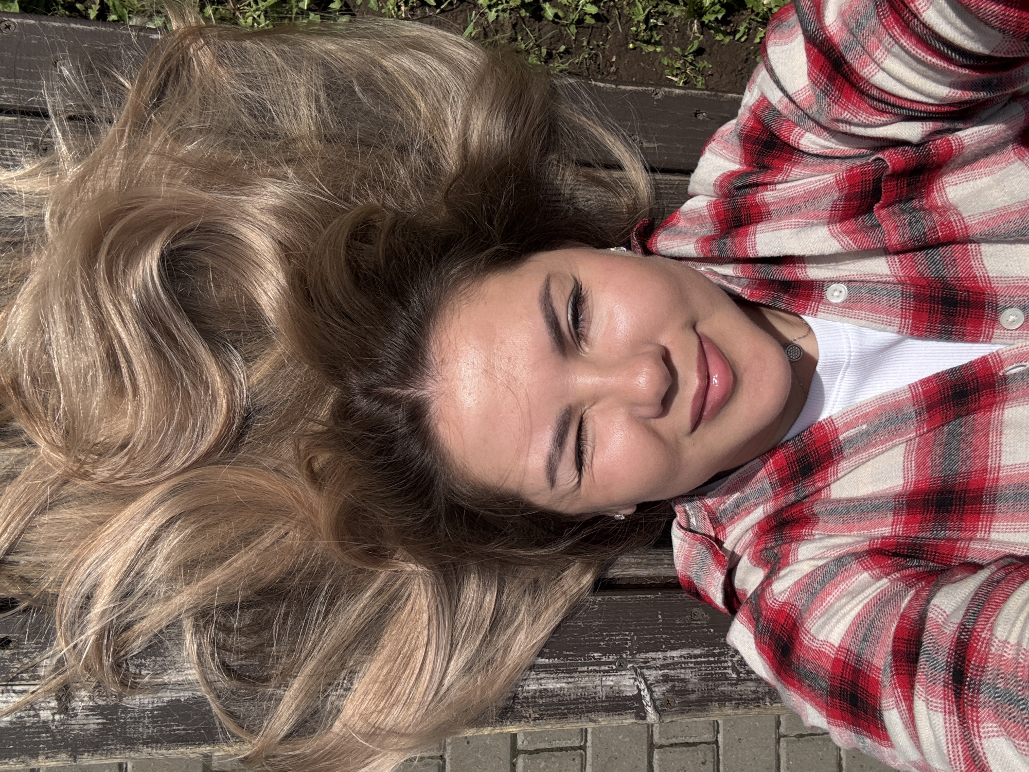 A young woman lies on a wooden bench outdoors. She has long, blonde hair spread out around her head, and she is wearing a red and white plaid shirt over a white top. She is smiling with her eyes closed. The bench is located on a paved surface, possibly a sidewalk, with grass and plants visible in the background. The location is unspecified, and there is no visible indication of the city of Izhevsk, Russia.