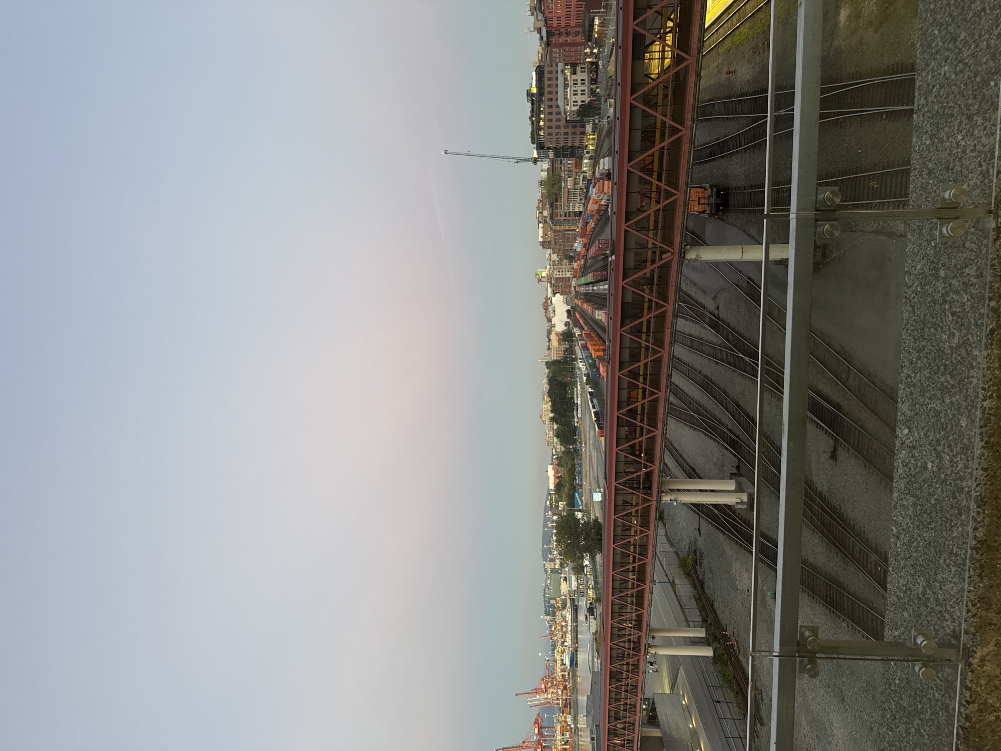 The image depicts a wide-angle view of a rail yard in the city of Vancouver, Canada, likely taken from an elevated vantage point. The scene is set at dusk or dawn, with a gradient sky displaying pale blues and pinks. In the foreground, a modern glass and metal railing overlooks several parallel sets of train tracks laid on a gravel bed.
Dominating the mid-ground is a long, elevated pedestrian bridge with a distinctive red, truss-like structure and clear paneling. This bridge spans across the tracks. Train cars, many of them stacked with shipping containers in orange and blue hues, are visible on the tracks beneath and beyond the bridge, suggesting active rail operations.
In the background, the Vancouver skyline is visible, featuring a dense collection of buildings, including industrial structures and more modern high-rises. To the left, the distinctive red cranes of a port facility are prominent, indicating the proximity of the shipping industry. A construction crane is also visible rising above the buildings in the distance. The overall impression is one of an active urban industrial area transitioning between day and night.