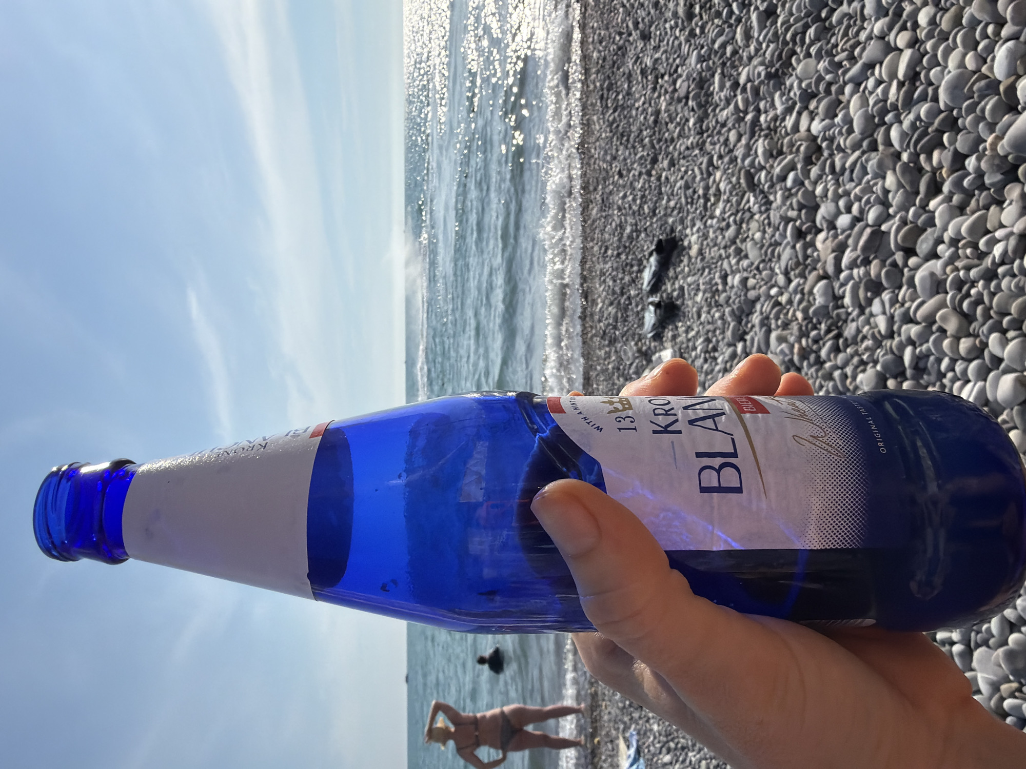 A first-person perspective shot shows a person holding a blue glass bottle of Kronenbourg 1664 Blanc beer at a pebble beach in Sochi, Russia. The beach has a gray pebble surface and is situated next to the Black Sea. In the background, the sea is calm with small waves. Two people are visible in the water; one woman is standing near the shore, wearing a bikini and a straw hat, and another person is partially submerged. The sky is clear and blue with scattered white clouds. The bottle's label indicates it is "ORIGINAL TASTE." The overall impression is a sunny day at the beach.