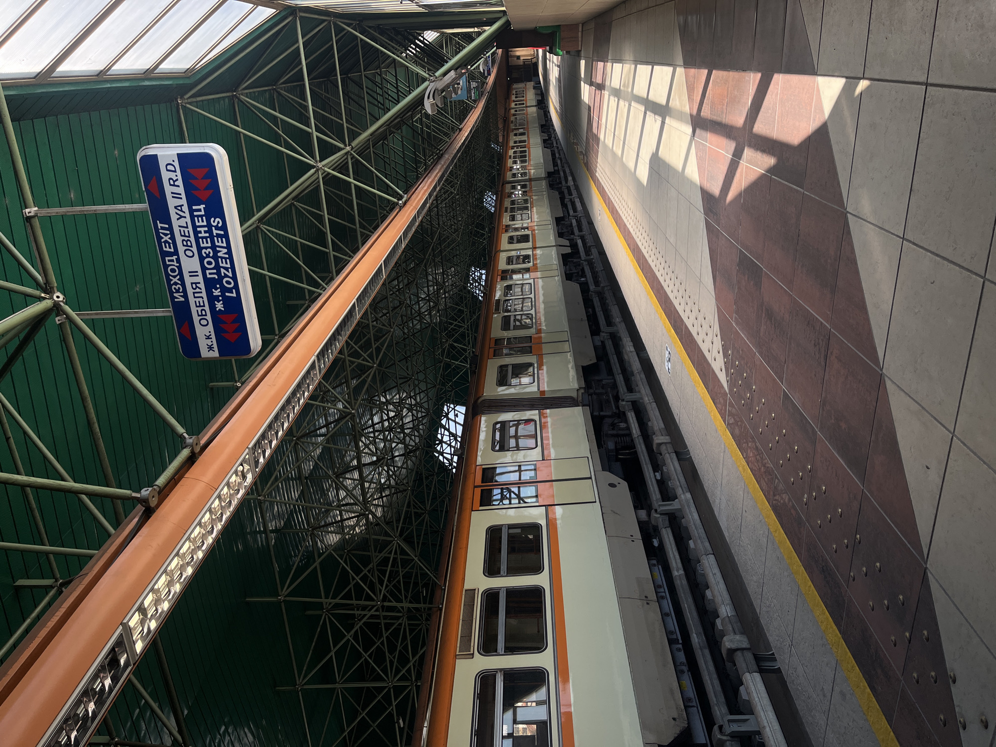 The image shows a subway station in Sofia, Bulgaria. A train with beige and orange colors is stopped at the platform. The platform has gray and reddish-brown tiling. Above the train and platform, there is a green-colored ceiling with a metal framework. A blue and white sign indicates "EXIT" in Bulgarian and English, along with the names of local districts in Sofia. The image is taken from a slightly elevated perspective, looking along the length of the train and platform. Sunlight streams through openings in the station's roof, casting shadows on the platform tiles. There are no people visible in the image.