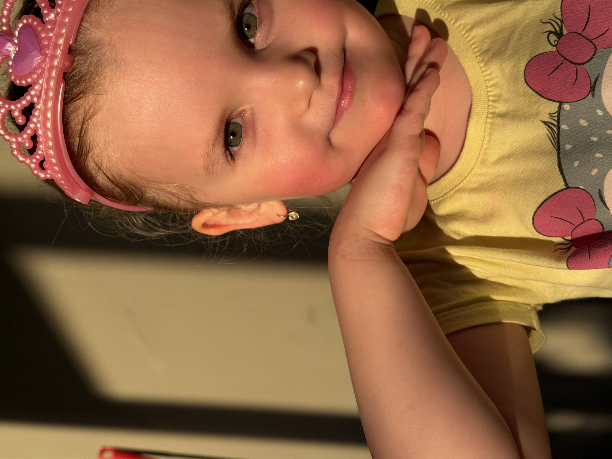 A young child with a pink tiara on their head is posing for a photo. The child has fair skin, rosy cheeks, and hazel eyes. They are wearing a yellow t-shirt with a cartoon character printed on it. The child's right hand is touching their face. The location is not identifiable as the city of Chaikovsky, Russia. The background is a light tan color with a dark shadow across the lower left of the image.
