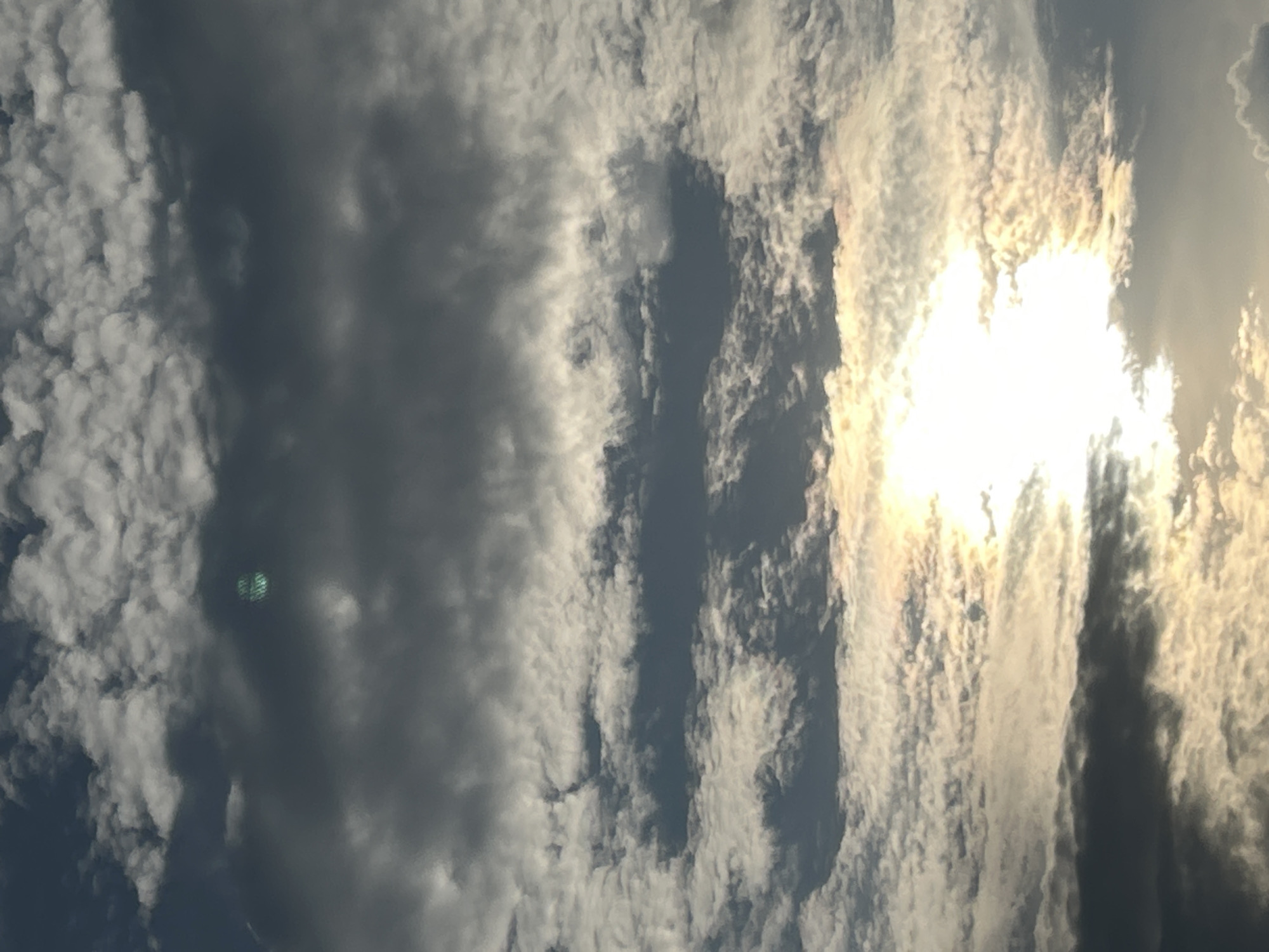 This is a photograph of the sky over Zelenograd, Russia, captured during daylight hours. The image is dominated by a dramatic display of clouds, with varying densities and textures. The lower half of the frame features a bright, diffused sunlight breaking through thinner, wispy clouds, creating a luminous glow. Above this, thicker, darker clouds cast shadows, suggesting a dynamic weather system. In the upper portion of the image, a light green, hexagonal lens flare is visible within a dark cloud formation. There are no people, structures, or ground-level details visible in this image; it is solely focused on the atmospheric conditions.