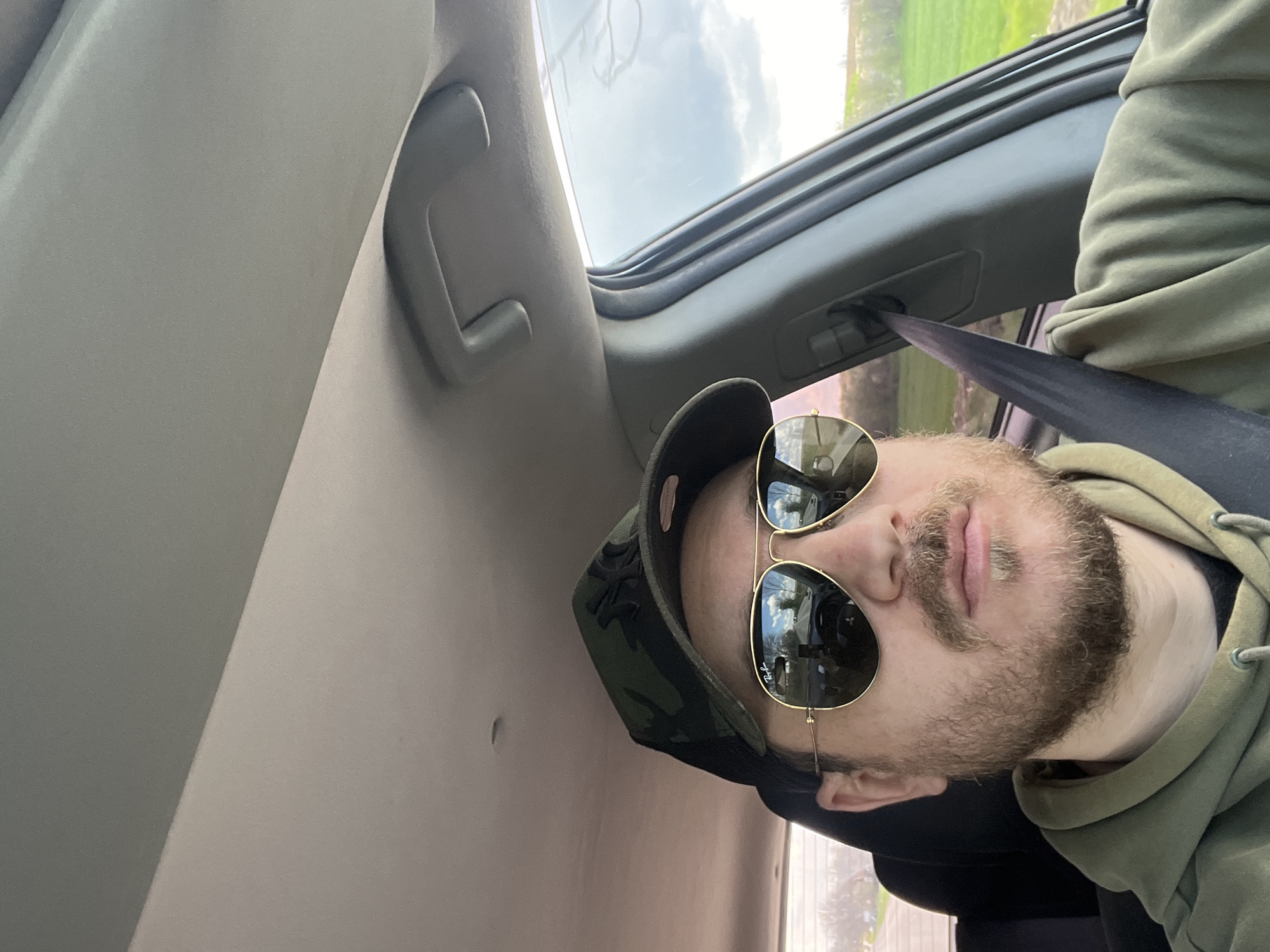 A man with a beard and sunglasses wearing a camouflage baseball cap and a green hoodie is in a car, wearing a seatbelt. The car has a sunroof and a gray interior. The location in the city of Ioglav, Bulgaria is not discernible.