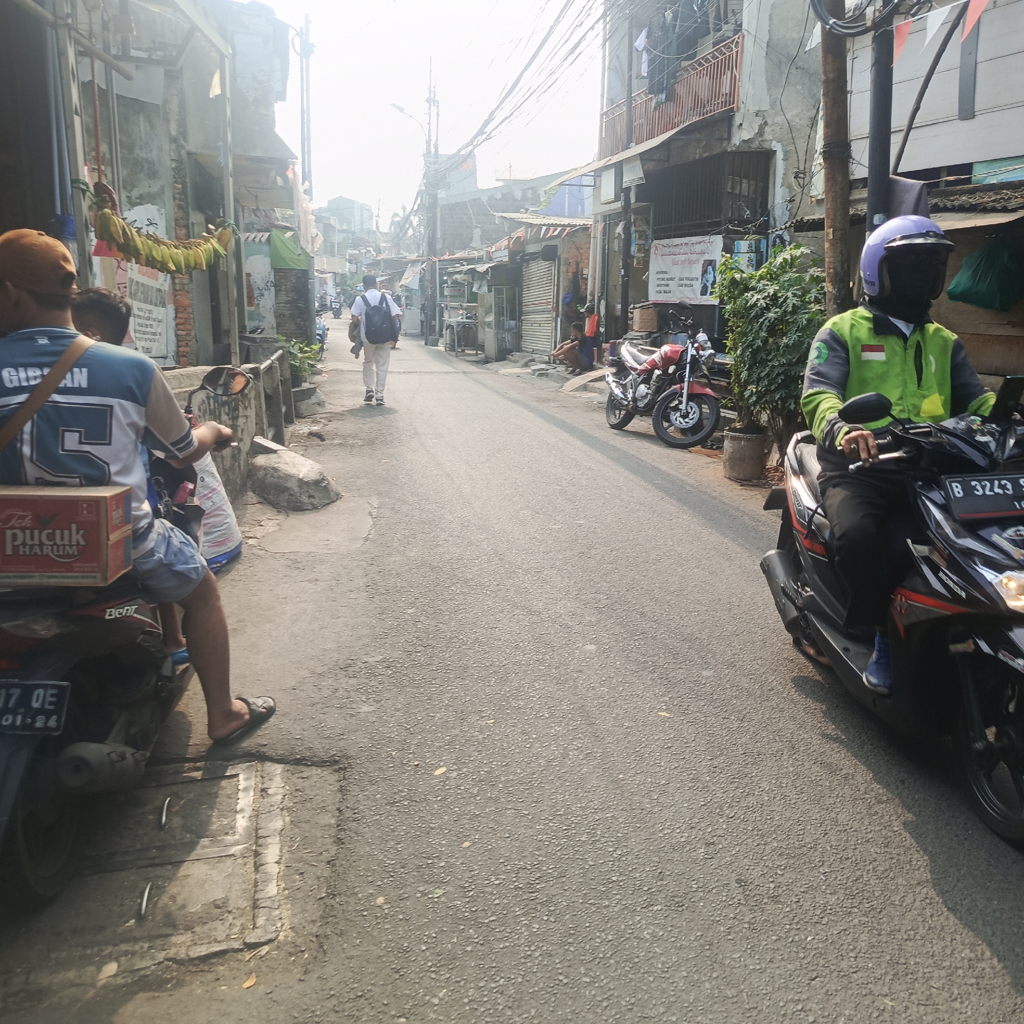 The image depicts a street scene in South Jakarta, Indonesia, under a bright, hazy sky. The street is paved and appears to be a narrow lane with buildings lining both sides. On the left, the buildings are characterized by weathered facades, some with exposed brick and corrugated metal roofs. Banana bunches are seen hanging from an overhead structure, possibly a market stall. Several individuals are present. In the foreground, a person on a scooter is partially visible, wearing a blue and white jersey and denim shorts, with a box of "Tek Pucuk Harum" tea strapped to the back of the scooter. Further down the street, a person in a white shirt and light-colored pants, carrying a black backpack, walks away from the camera. On the right side of the street, a person wearing a green and grey uniform with a purple helmet, riding a black scooter, is prominent. The scooter has a license plate that reads "B 3243 S". More buildings, some with signs including one for "KREASI SALON", are visible along this side, interspersed with trees and other vegetation. Several other scooters are parked or in motion further down the street, indicating a typical urban environment. The overall impression is a bustling street in a dense urban area.