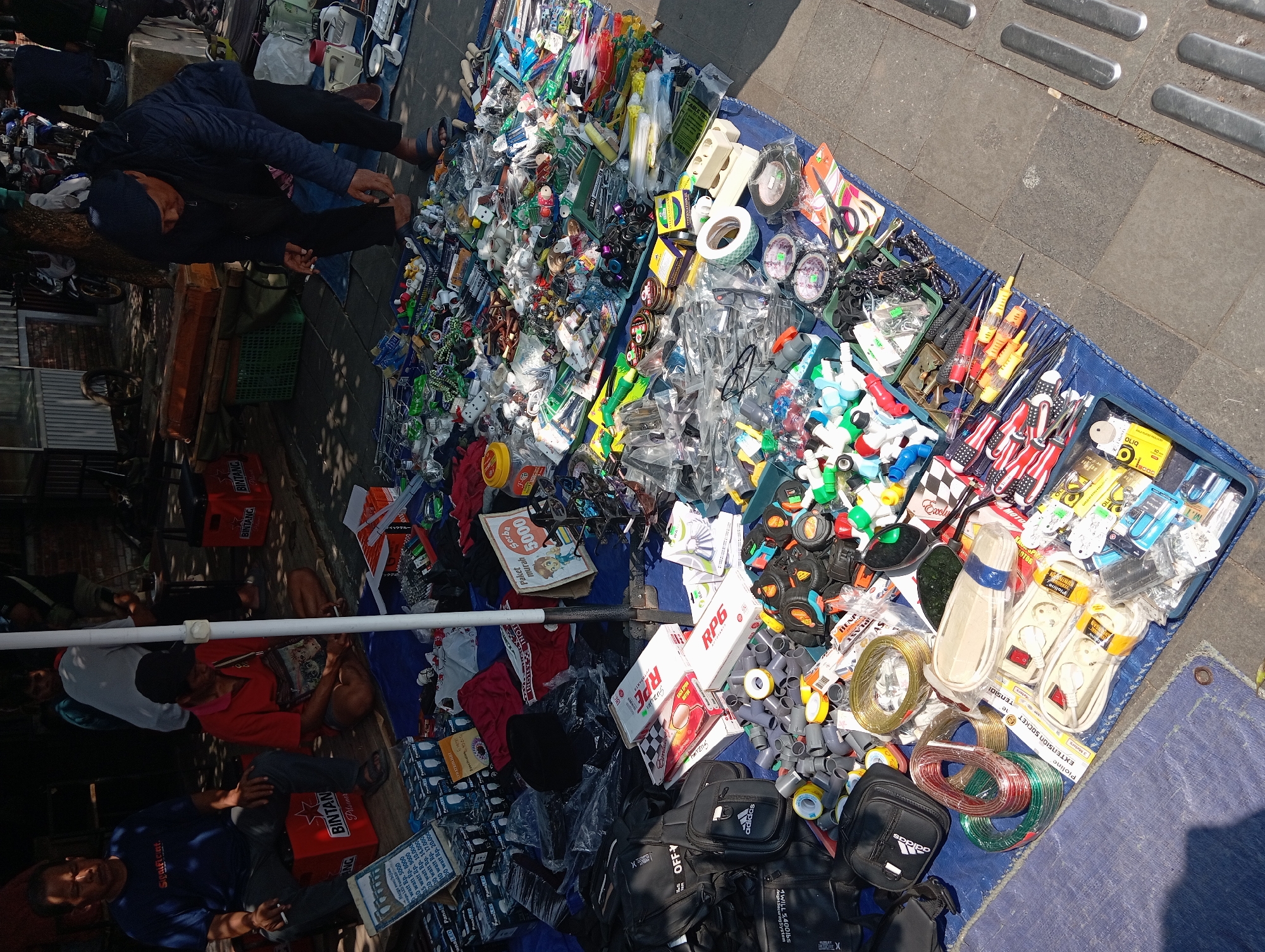 In Borobudur, Indonesia, on a sunlit day, a street market is set up on a blue tarpaulin spread across a tiled sidewalk. Several individuals are present, some attending to the merchandise, others observing or interacting. In the foreground, a variety of items are displayed, including an assortment of electrical extension cords, rolls of wire in different colors, a box of fuses, and several packaged electrical outlets. To the left, a man in a blue t-shirt sits on an orange crate, smoking a cigarette. Beside him, another man in a red shirt is seated on the ground, also smoking, with a bag slung over his shoulder. Further back, a person in a dark jacket and cap is bent over, examining items on display. The market is filled with numerous small objects, many of which appear to be hardware or electronic components, such as screws, connectors, and small tools. The backdrop includes some market stalls and what appears to be a tent structure. The overall scene depicts a bustling outdoor market environment.