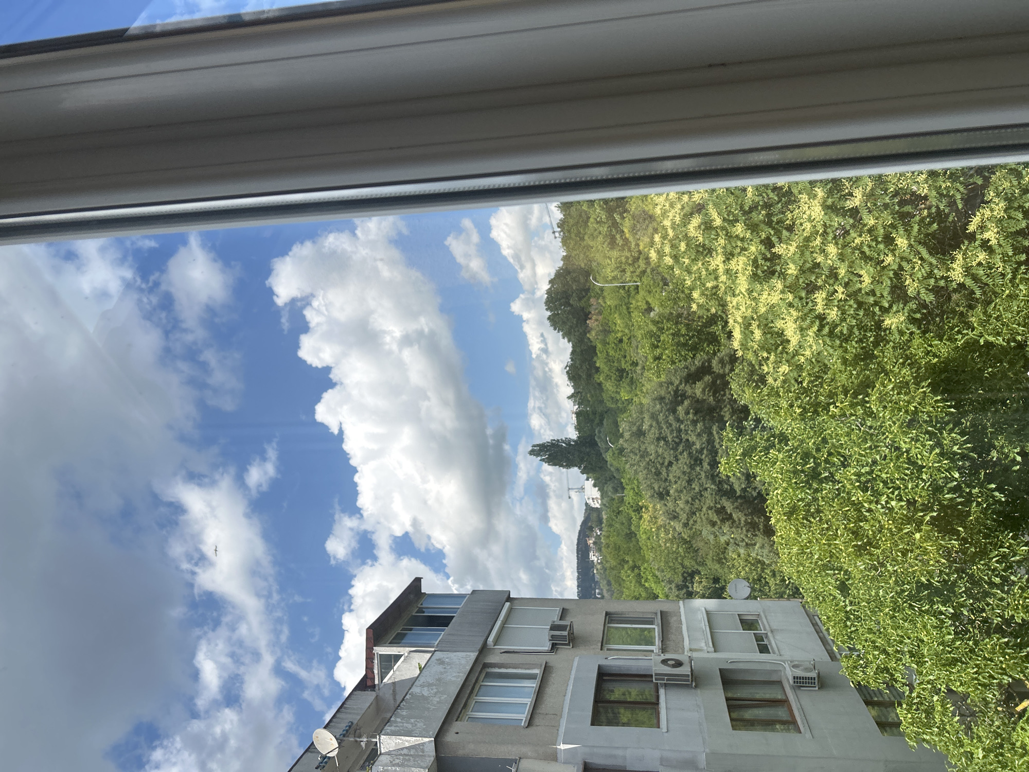 This is a vertical, eye-level, outdoor shot looking out from a window frame. The top of the image is dominated by the white and grey frame of the window, with a hint of the sky visible through it. Below the window frame, the scene opens up to a view of a blue sky filled with fluffy white clouds. A solitary bird is visible in the sky.
In the middle ground, a grey, multi-story apartment building with multiple windows and air conditioning units is partially visible. One of the apartment building's balconies has satellite dishes. Behind the building, a dense expanse of lush green trees, some with lighter, yellowish foliage, covers the landscape. The trees are abundant and create a wall of green. In the distance, faint hints of other buildings or structures can be seen through the trees, suggesting an urban or semi-urban environment. The overall impression is a bright, sunny day, likely in the late morning or early afternoon, given the quality of the light. The scene appears to be in Varna, Bulgaria, as indicated by the location context. No specific activity is taking place, it's a static view of a residential area with natural elements.