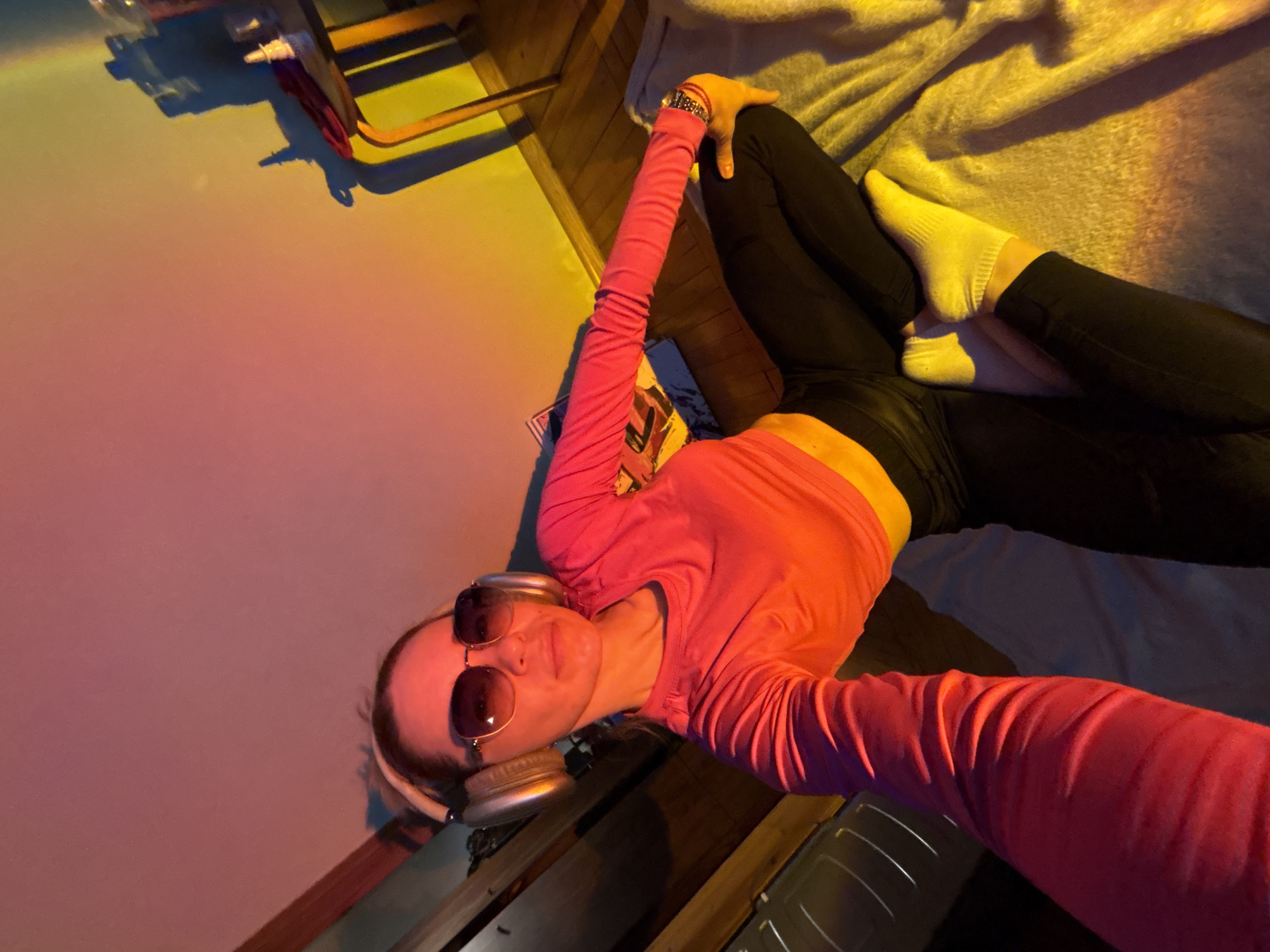 A person is laying down on a bed with a white sheet. They are wearing black leggings, a pink long-sleeved shirt, and sunglasses. They are also wearing headphones around their neck. The person is facing the camera. The room is lit with a yellow and pink light.
