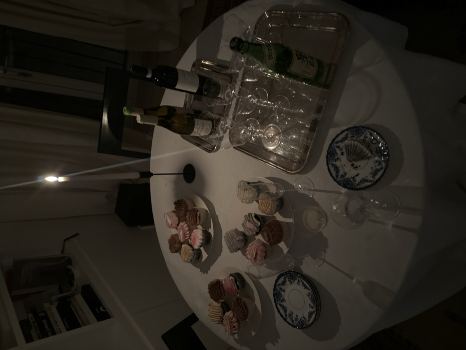 The image depicts an indoor dining scene at night, illuminated primarily by a single lit white candle in a black candlestick, casting prominent shadows. The central subject is a round table draped with a white tablecloth, located within a private residence in London, United Kingdom. On the table, three white plates hold an assortment of approximately 25-30 cupcakes, featuring various colored frostings such as pink, brown, white, and tan. Two silver trays are present: a smaller tray contains one bottle of white wine and one bottle of red wine, along with two empty stemmed glasses. A larger rectangular silver tray holds six empty stemmed wine glasses and one green bottle with Japanese characters. Several additional empty stemmed glasses are placed directly on the tablecloth. A stack of three blue and white patterned dessert plates is visible, with one individual plate holding a discarded cupcake wrapper. The background shows a white wall unit with books to the left and a dark-backed chair followed by dark curtains or drapes to the right. No individuals are visible in the scene.