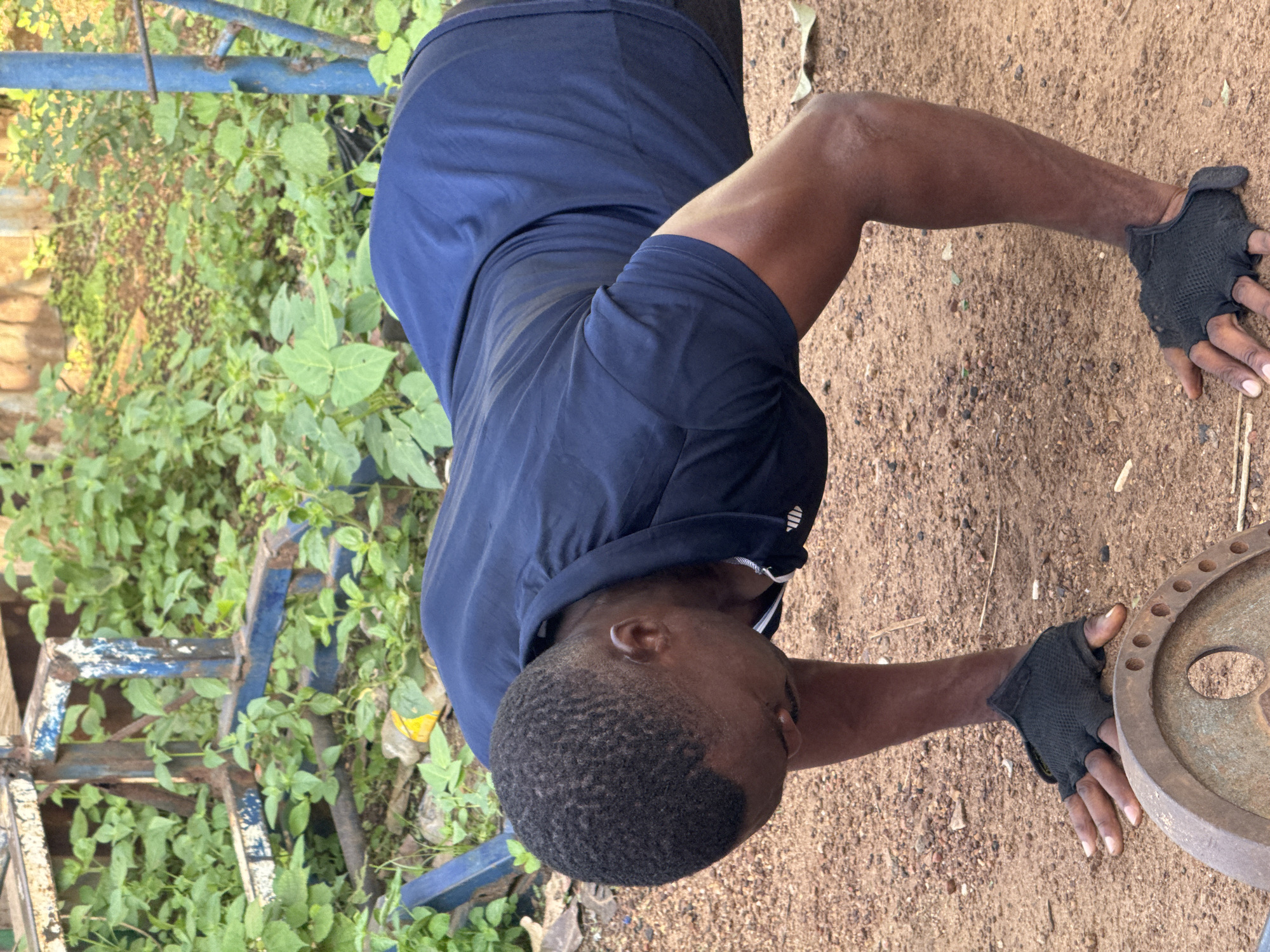 In Akwanga, Nigeria, a Black male is performing push-ups outdoors. He is wearing a dark blue t-shirt and black fingerless gloves. His hands are on the ground, with a rusty weight plate visible in the foreground. The background consists of overgrown green vegetation and rusty metal gym equipment. The ground appears to be dirt and gravel.