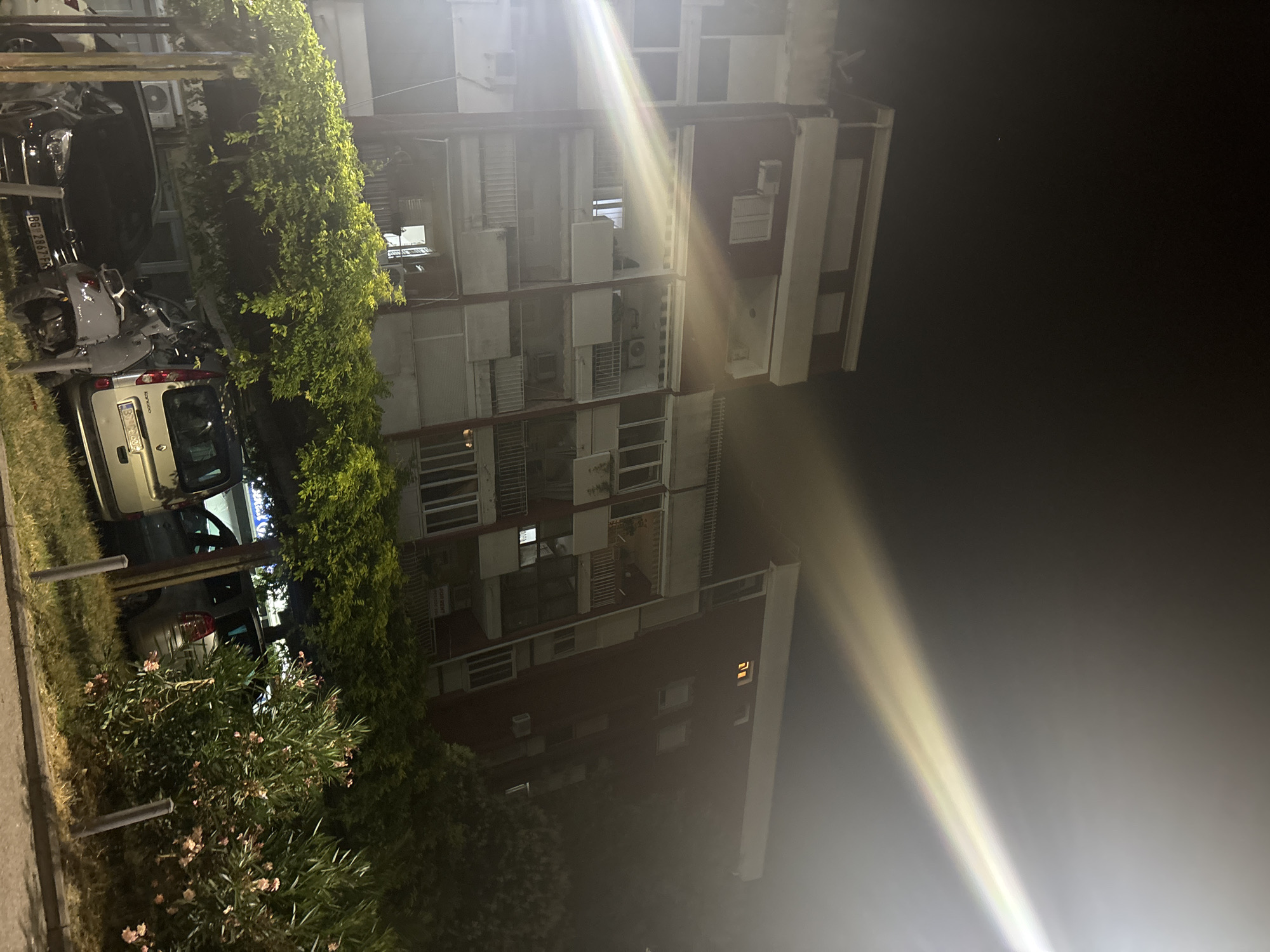 The image depicts a nighttime scene in Budva, Montenegro, focusing on a multi-story apartment building. A bright light source, possibly a streetlamp, casts a strong beam across the upper right portion of the frame, creating a lens flare effect. The building, a typical example of mid-20th-century architecture, has multiple balconies and windows, some of which are illuminated, suggesting occupancy. In the foreground, there is a row of parked cars and a scooter beneath a canopy of lush green foliage. The overall atmosphere is urban and residential, characteristic of a city at night.