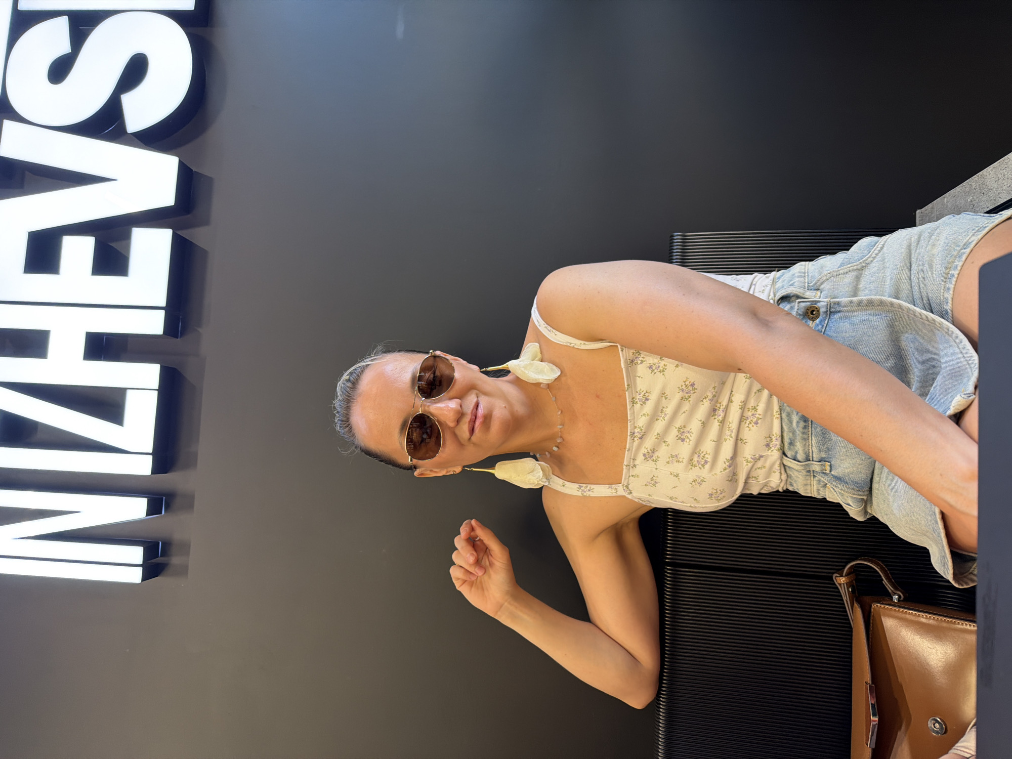 A woman with blonde hair pulled back is sitting on a black bench in front of a dark wall with illuminated white letters spelling "IZHEVSK". She is wearing a floral print tank top, denim shorts, large aviator sunglasses, and dangling earrings. Her left arm is resting on the back of the bench, and her right hand is raised slightly. A brown leather purse is on the bench to her left. The lighting suggests an indoor environment, possibly a store or public space in Izhevsk, Russia.