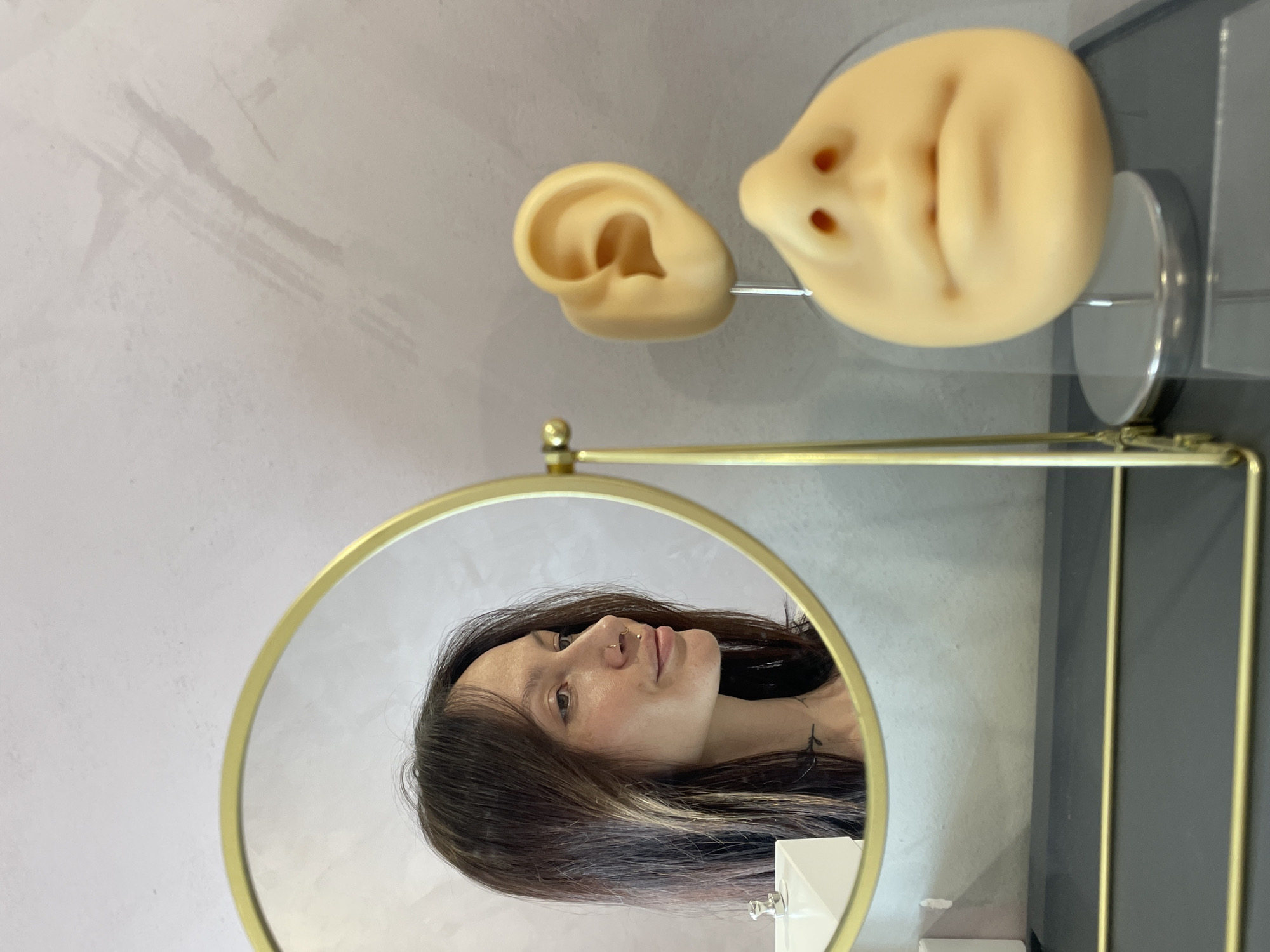 The image shows a woman's face reflected in a gold-framed, round mirror. Above and to the right of the mirror are two prosthetic body parts, one resembling an ear and the other resembling a nose. The setting appears to be indoors, possibly a medical clinic. The location in Izhevsk, Russia, is not determinable from the image.