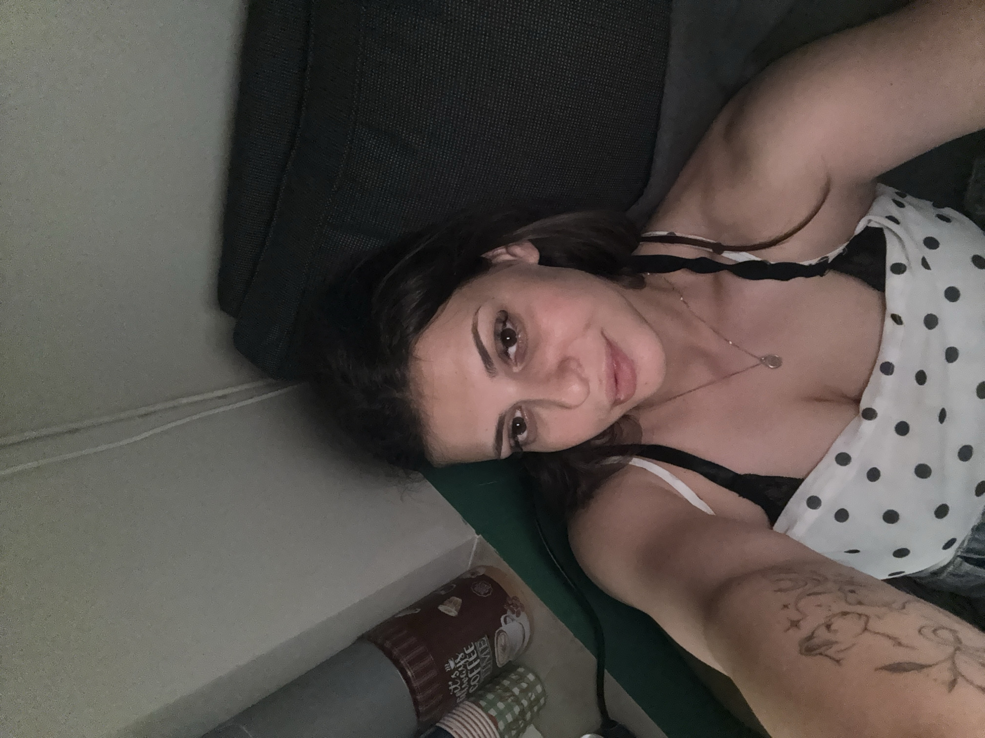 A young woman with dark hair, wearing a white polka dot top and a black bra, is lying down on a dark grey couch. She is looking at the camera with a slight smile. Her left arm, visible on the left side of the frame, has a tattoo. Behind her, on the left, is a green surface, possibly a yoga mat or a rolled-up mat, next to a container labeled "Always Coffee Time" and some stacked paper cups. The background is a plain, light-colored wall. The scene appears to be indoors, in what could be a living room or bedroom. There is no discernible information within the image to place the location specifically in the city of Plovdiv, Bulgaria.