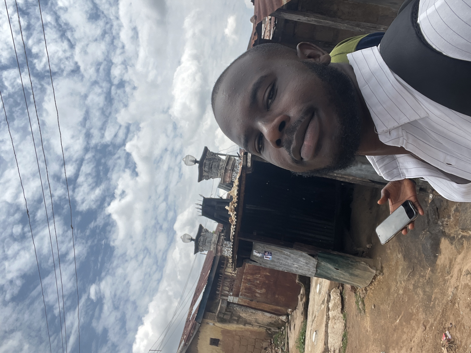 A male individual is depicted taking a selfie outdoors in Jos, Nigeria, under a cloudy sky. The subject, a man with dark skin, a shaved head, and a full beard, is positioned in the foreground, looking towards the camera with a neutral expression. He wears a white collared shirt with thin vertical stripes and a black backpack strap over his right shoulder. In his left hand, he holds a silver-colored smartphone, angled for the selfie.
The background reveals a semi-urban environment characterized by older, modest structures. To the left, a building with mud-colored walls and a rusted corrugated metal roof is visible, featuring a small square window. In front of this, a corrugated metal shack or shed stands with a dark green base. Dominating the midground skyline are two decorative concrete pillars, each topped with a light fixture, situated on the roofline of a building beyond the shacks. The ground is unpaved earth, with visible concrete sections and scattered debris. Three dark power lines extend horizontally across the upper portion of the frame, set against a sky filled with white and grey clouds. A partial view of another person's shoulder, wearing green and dark blue clothing, is visible behind the main subject on the right.
