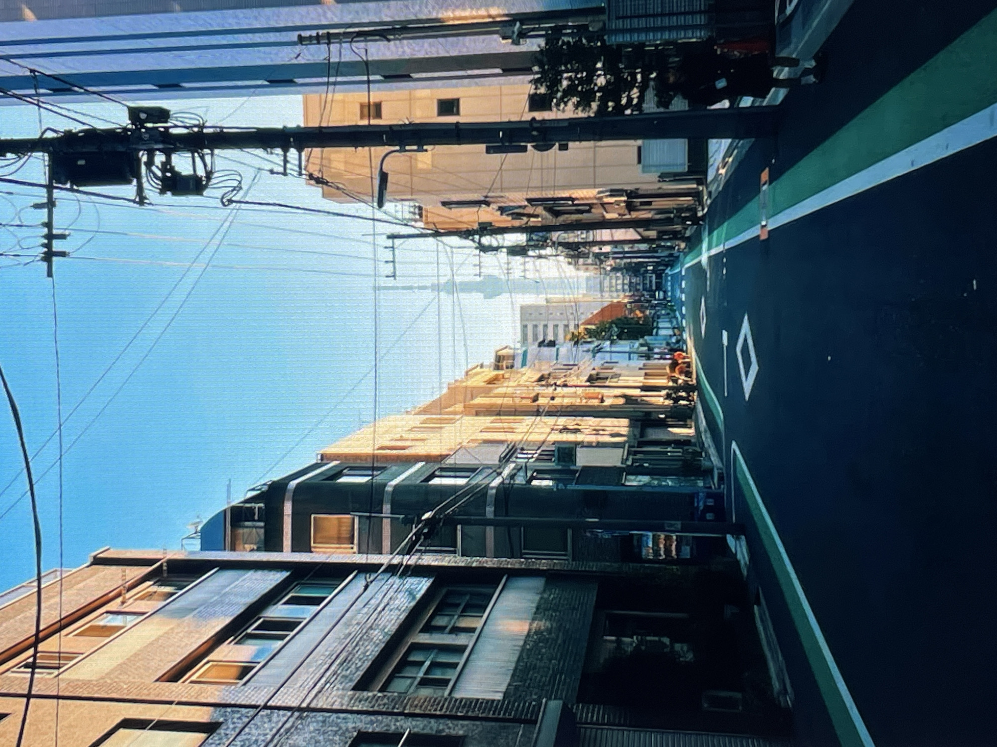 The image depicts a city street in an urban environment, possibly in Japan, given the architecture and power lines. The perspective is from street level, looking down the avenue. Buildings line both sides of the road, varying in height and architectural style. A distinct road marking indicates a lane division, and numerous overhead wires crisscross the sky, supported by utility poles. In the distance, a tall structure, possibly a communication tower, is visible. The sky is clear and blue.