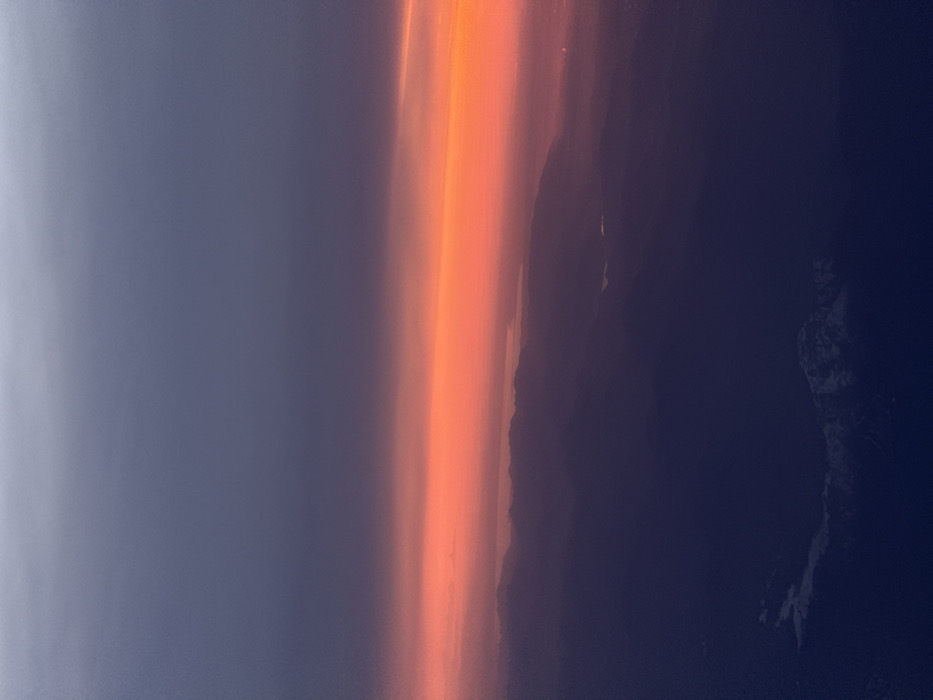 An aerial panoramic view captures a dramatic sunset or sunrise over a mountainous landscape, consistent with a perspective overlooking regions near Mariano Comense, Italy. The sky presents a gradient, transitioning from a deep, dusky gray-purple at the top to an intense horizontal band of vibrant orange and red across the middle, brightest towards the right. Below this luminous horizon, multiple layers of dark, silhouetted mountain ranges extend into the foreground, their forms indistinct due to distance and atmospheric haze. Several calm bodies of water, likely lakes, are interspersed within the valleys, reflecting minimal light. Patches of white, indicative of snow, are visible on the peaks of the lower, more defined mountains in the foreground, particularly towards the bottom left and right. No specific human activity, structures, or individuals are discernible in the natural environment, though a faint glow in the far right distance suggests distant settlements.