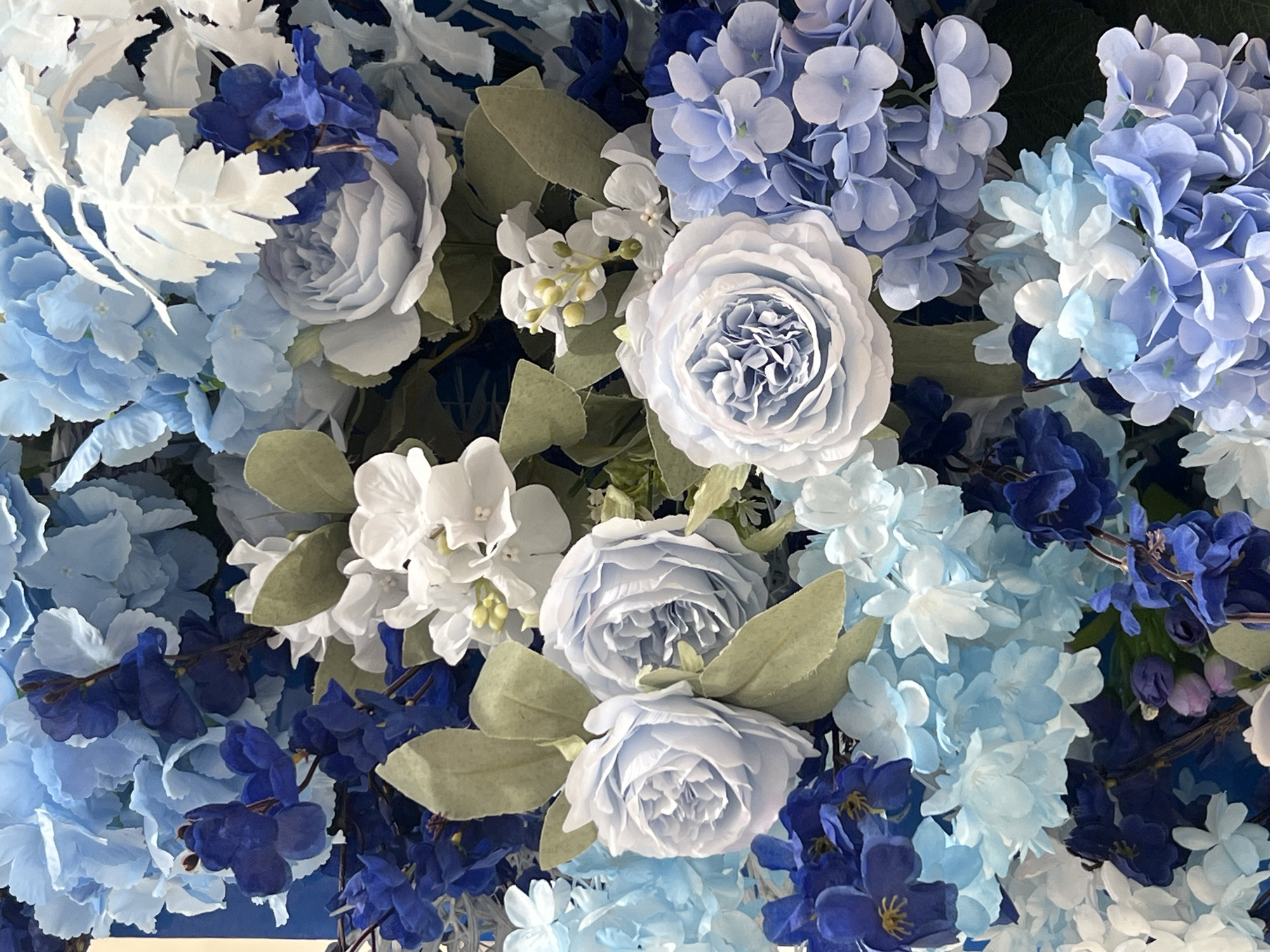 This is a close-up, overhead, eye-level shot of a dense floral arrangement dominated by various shades of blue and white flowers, interspersed with green leaves and some darker blue foliage. The flowers are arranged tightly, creating a textured and layered effect. Prominent are large, light blue roses with intricate petal formations, resembling peonies, and clusters of smaller, round blue hydrangeas. There are also clusters of delicate white flowers and deep blue, almost black, flowers with elongated petals. The overall impression is of a lush, artificial floral display. There is no indication of any specific location within the city of Lisky, Ukraine, nor are there any people, actions, or interactions depicted. The scene is static and purely decorative.