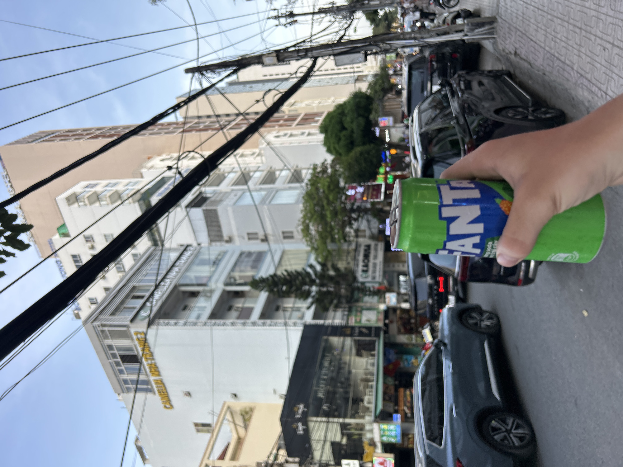 A person's hand holds a green can of Fanta soda in the foreground, obscuring a portion of a black SUV and the right side of the image. The background depicts a street scene in Nha Trang Ward, Vietnam, with buildings of varying heights lining the street, a tall, slender tower visible amongst them. Numerous electrical wires crisscross the sky above. The street is occupied by several vehicles, including cars and motorcycles. A tiled sidewalk runs along the right side of the street, with a utility pole and more overhead wires present. The overall impression is of a bustling urban environment.