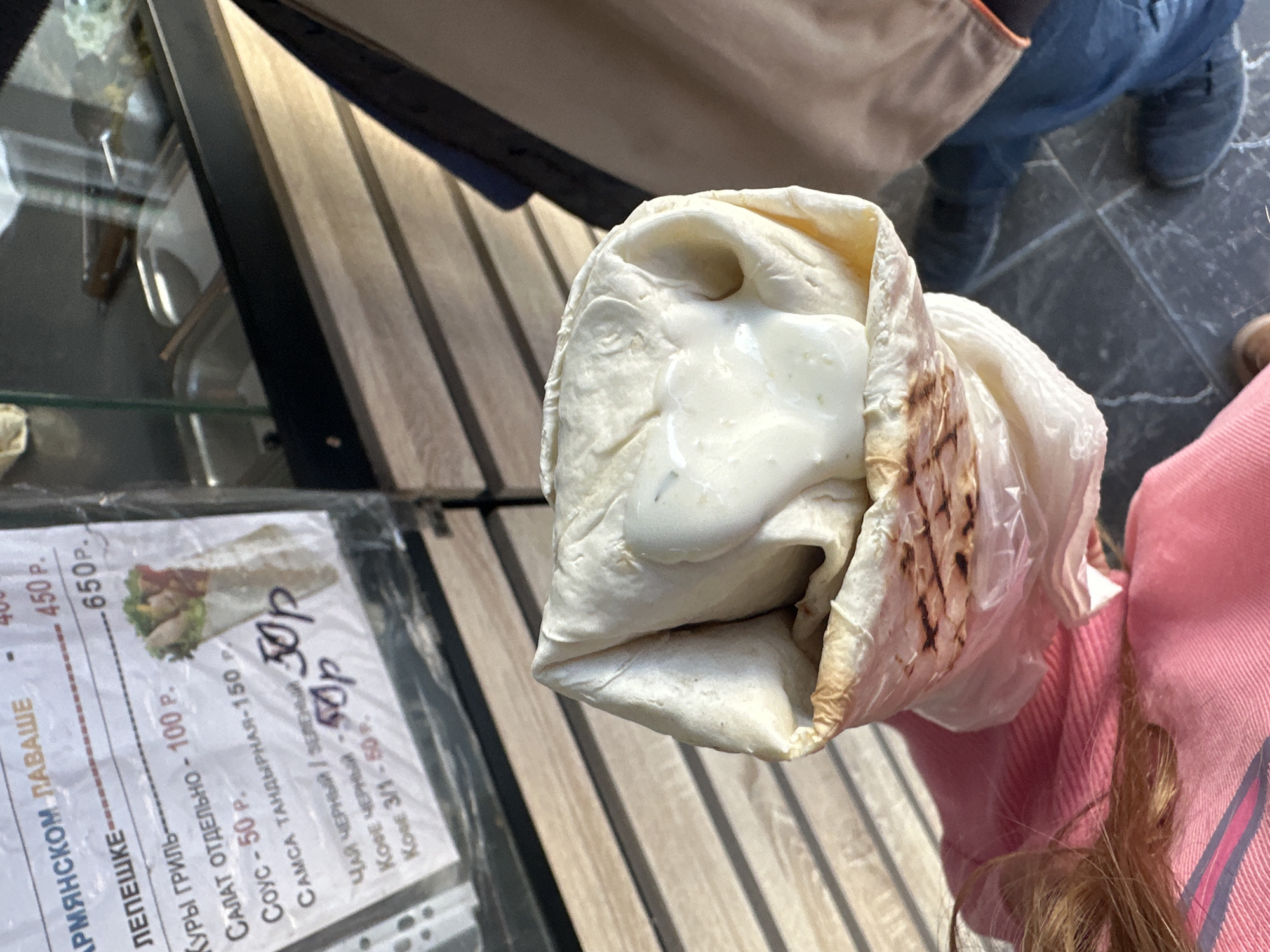 A shawarma wrap with white sauce oozing out is held in front of a menu board advertising “Armenian Lavash”, or shawarma, in the Russian language. The menu also advertises the price of the sauce as 50₽. Parts of two people are visible - one wearing blue jeans and a dark shoe, and the other with long blond hair wearing a pink garment. This food stall is most likely located in the city of Moscow, Russia.