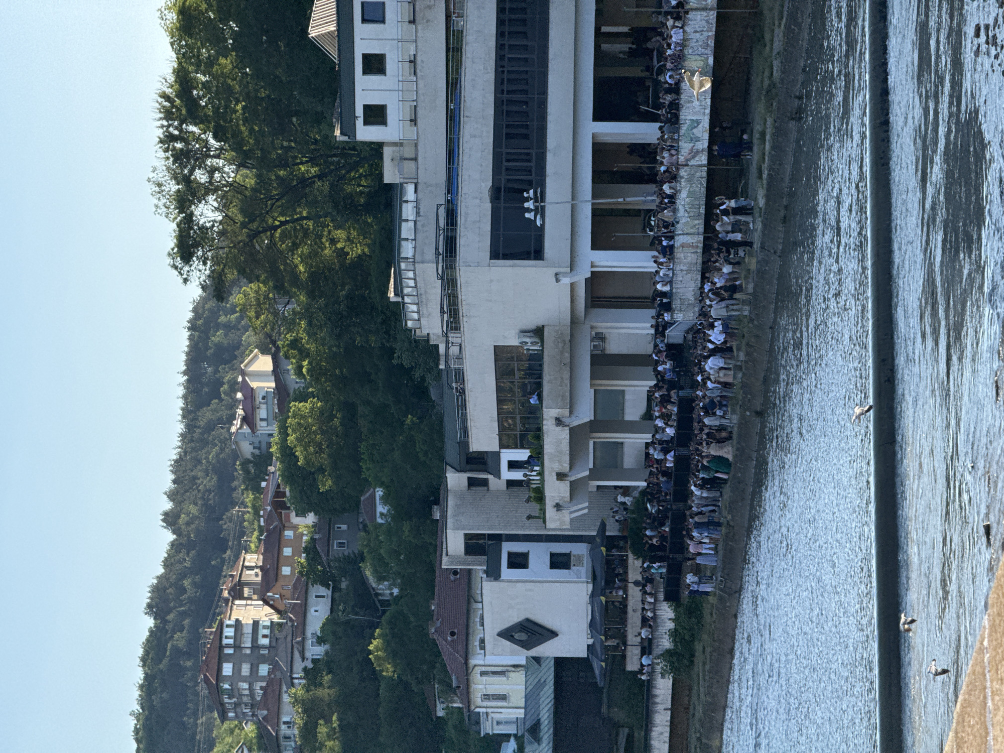 The image depicts a riverfront scene, with a building complex situated along the river's edge. A large crowd of people is gathered along the riverbank in front of the building. On the far side of the river is a hillside with houses and trees. The location appears to be a city.