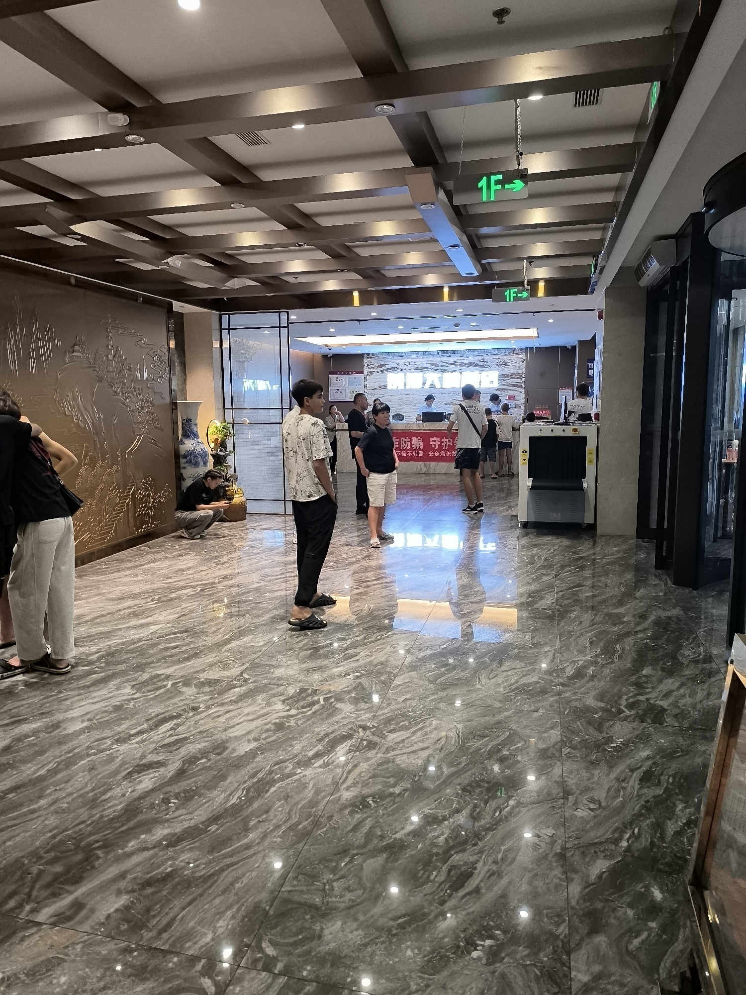 The image captures a well-lit indoor space, likely a lobby or waiting area, with a polished marble-patterned floor and a decorative ceiling featuring wooden beams. Several individuals are present, some standing and others seated. In the foreground, a person in a black top and light-colored pants stands facing away from the camera. To their left, another person in a similar outfit is also partially visible. Further into the scene, a young person wearing a patterned white shirt and black pants stands facing right. Another individual in a black t-shirt and shorts is seated on the floor near a large decorative vase and a plant.
In the mid-ground, there is a reception desk with several people interacting. A security scanner is positioned in front of the desk. Above, green illuminated signs with "1F" and arrows indicate the direction to the first floor. The overall environment suggests a public building, possibly a hotel or administrative center, in Ürümqi, China. The lighting is bright, casting reflections on the glossy floor. The walls are adorned with what appears to be a large, ornate relief carving.
