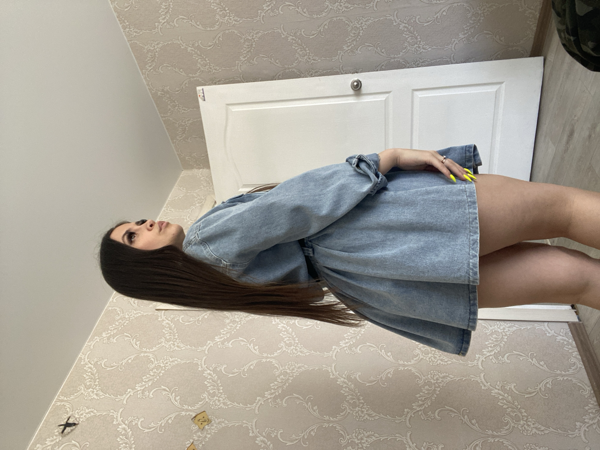 A woman with long brown hair is wearing a denim dress. She is standing indoors against a wall with floral wallpaper. In the background is a white door. The woman's nails are painted bright yellow. The location appears to be an interior space in the city of Chaikovsky, Russia based on the metadata.