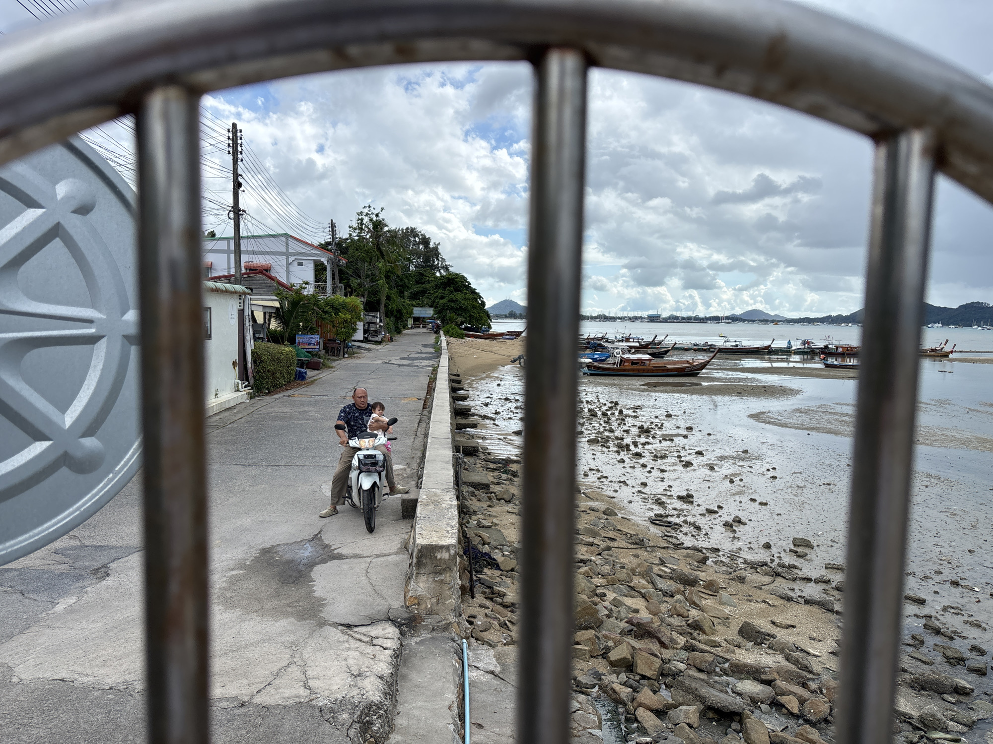 The image shows a view through metal bars of a street next to a beach in Rawai, Thailand. A man is riding a white motorbike with a child sitting in front of him, on the paved street. The street is lined with buildings and trees on the left, and a rocky beach with several longtail boats is on the right. The sky is cloudy. The overall scene depicts a coastal area with a mix of residential and maritime elements.