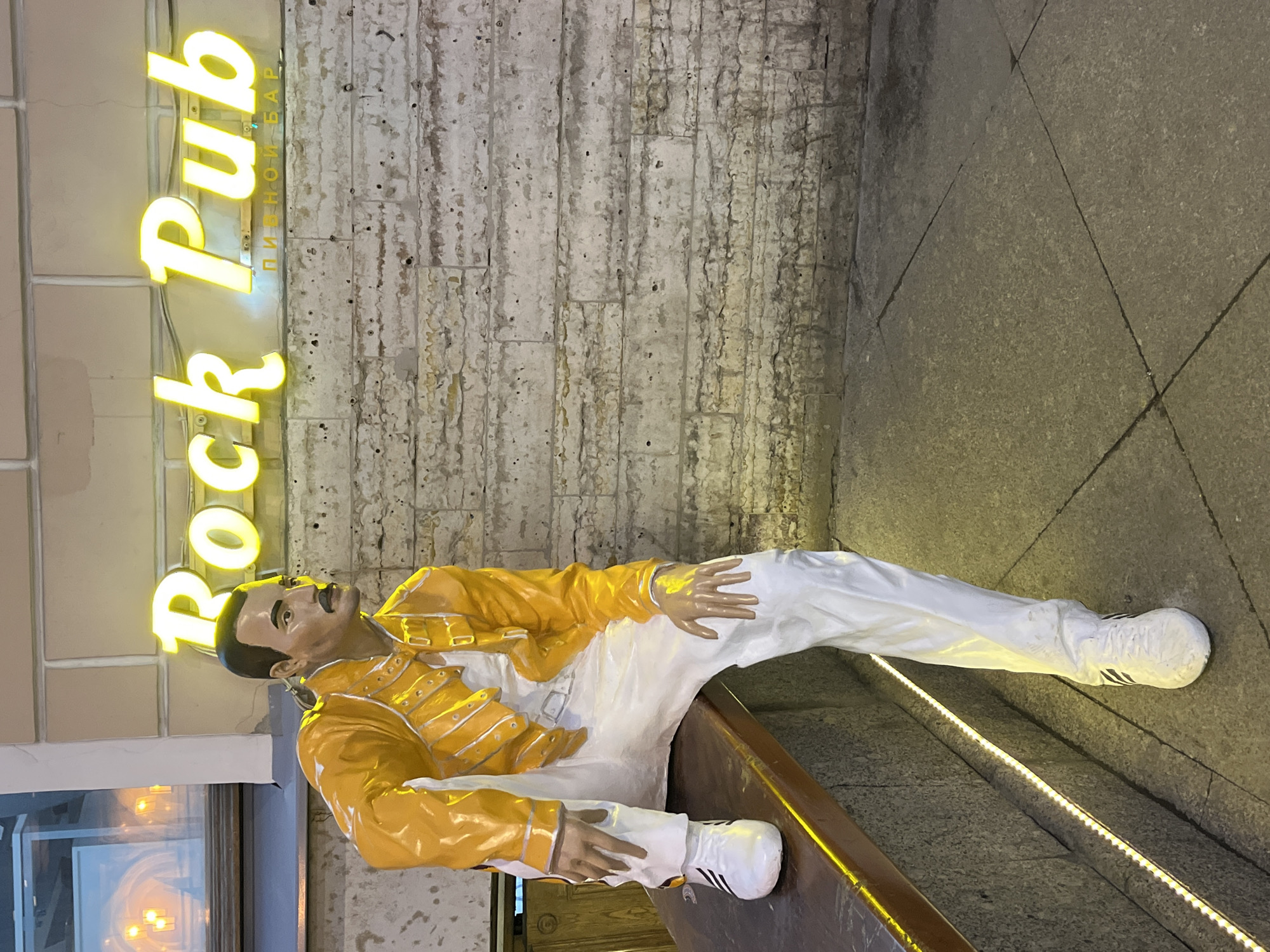 A statue of Freddie Mercury reclines below a neon sign reading "Rock Pub" in Saint Petersburg, Russia. Mercury wears his iconic yellow jacket and white pants, one foot rests on a surface. The background consists of a brick wall and a paved area.