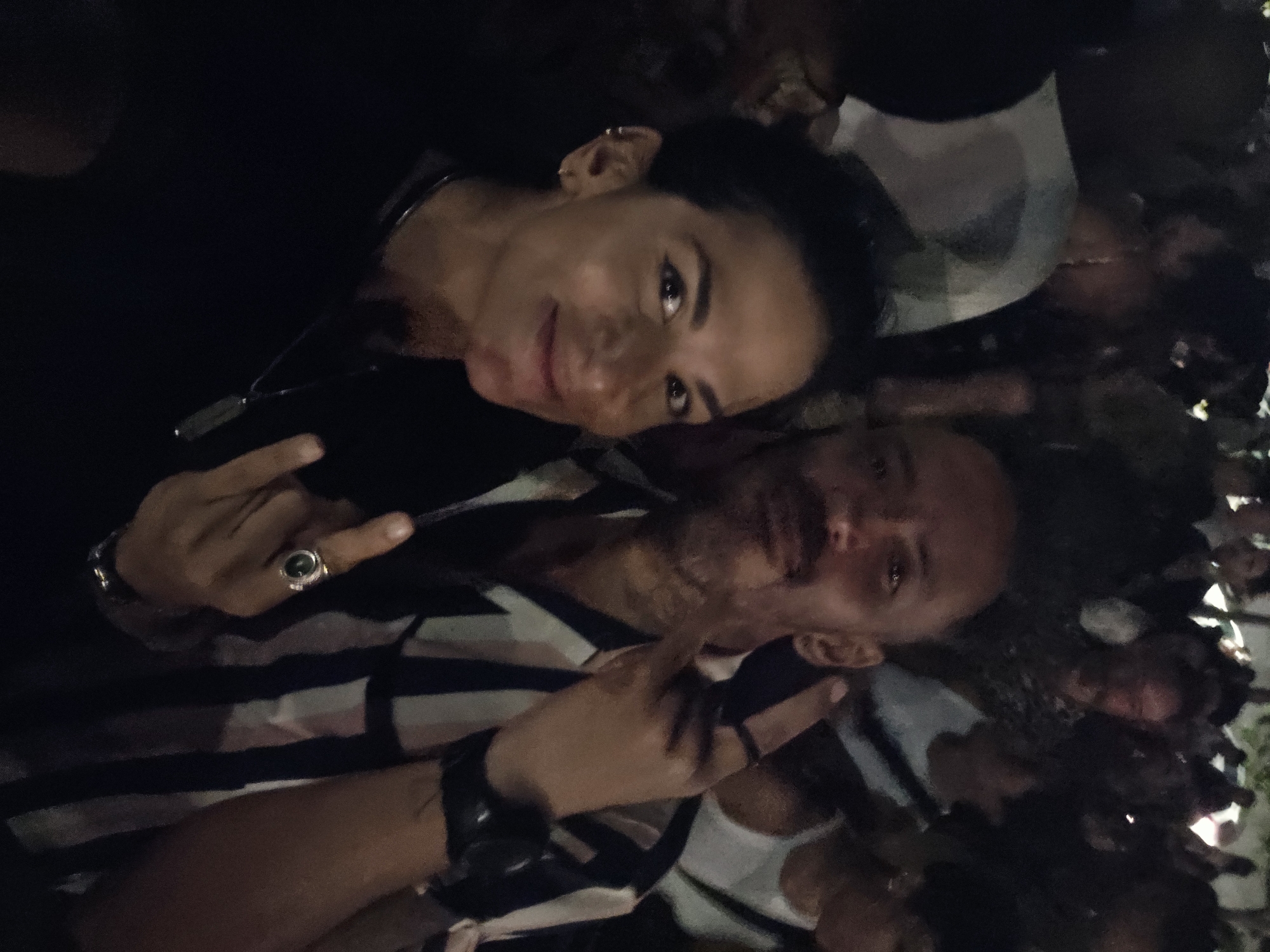 Two individuals pose for a photo in a dark, crowded setting. The woman on the left wears a black shirt and a necklace, with multiple earrings in her left ear. Her skin appears fair and her hair is dark. The man on the right wears a striped shirt, a watch on his left wrist, and a ring on his left hand. Both are making the "rock on" hand gesture. The background reveals a dense gathering of people, suggesting a public event. While the exact location within Armação dos Búzios, Brazil is unclear from the image, the crowded environment points towards a potentially popular venue or event space.