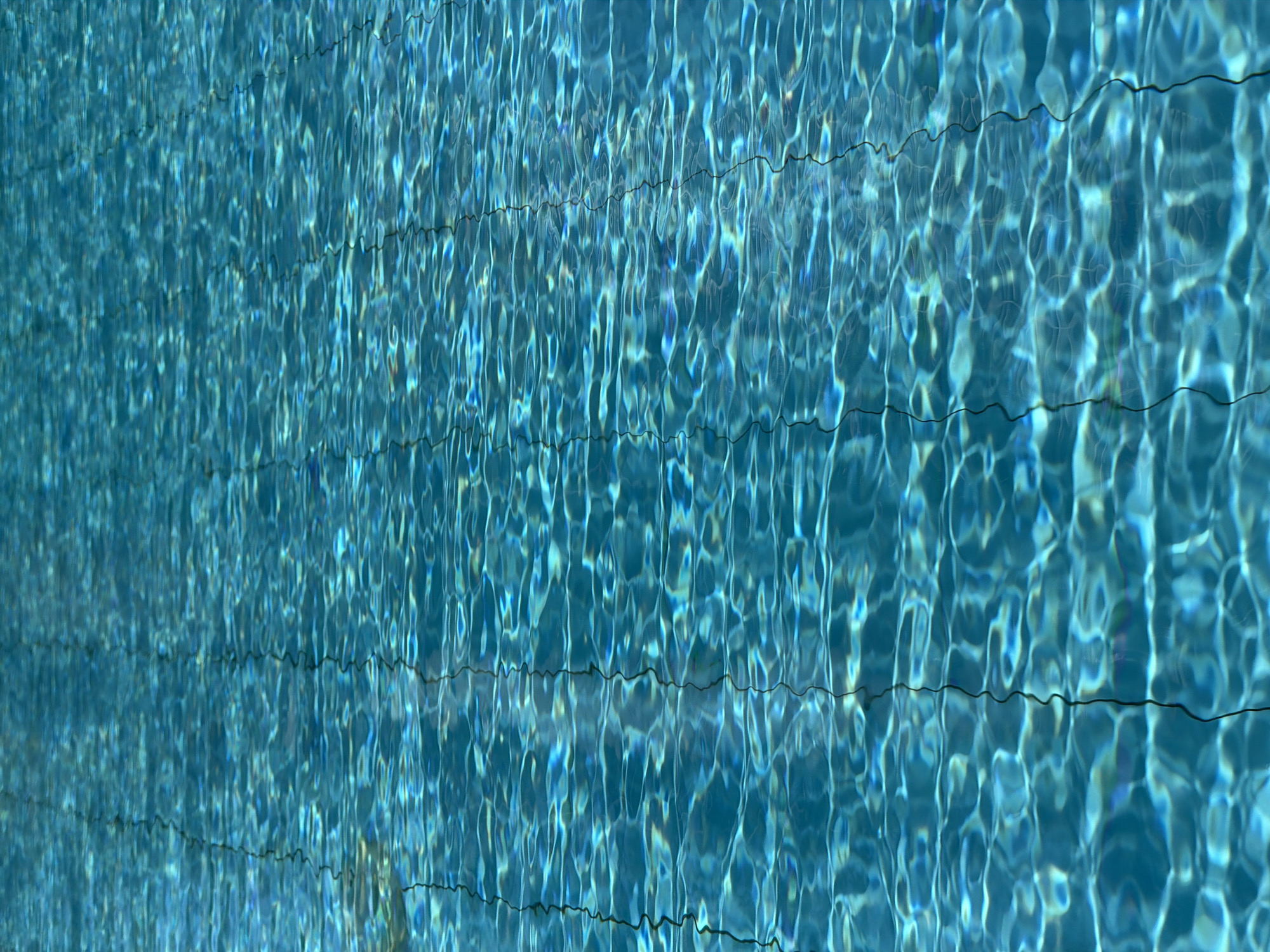 The image shows the surface of a swimming pool in Nha Trang Ward, Vietnam. The water is clear blue with wavy reflections and refractions. Horizontal lines of grout separate tiles beneath the water's surface. No people or other objects are visible.