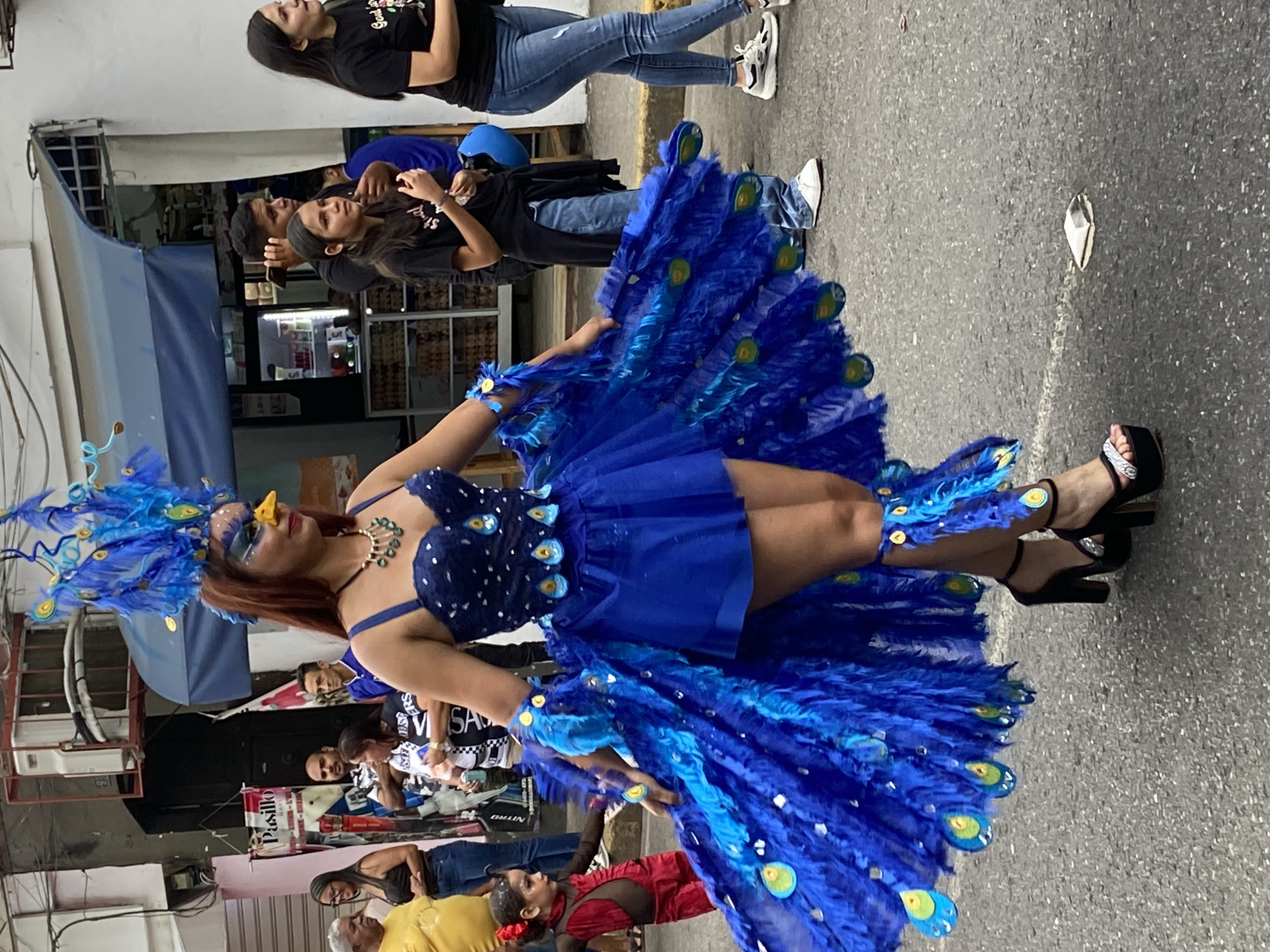 A street scene in Tinaquillo, Venezuela, shows a woman in an elaborate blue peacock-themed costume. The woman wears a blue dress with peacock feather embellishments, a feathered headdress, and high-heeled sandals. She is standing on a paved street surrounded by people, some of whom are watching her. Buildings, signs, and parked vehicles are visible in the background. Some other people are wearing street clothing.
