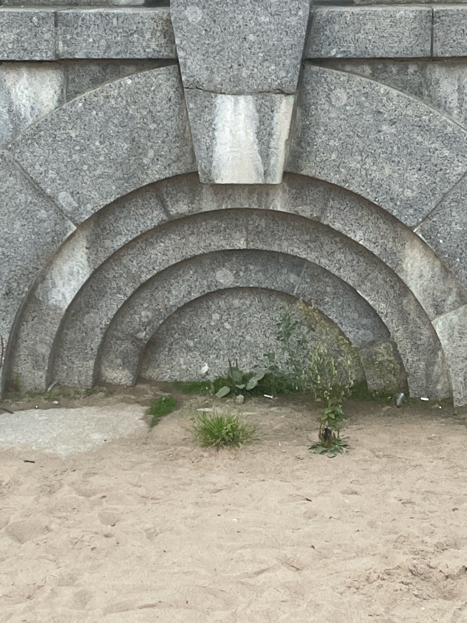The image shows a section of a granite wall with several concentric arches carved into it. The wall is constructed from rough-hewn granite blocks, and the arches are progressively smaller, creating a layered effect. The ground in front of the wall is covered in sand and sparse vegetation, including a small bush and some blades of grass. The scene appears to be outdoors, with natural light casting shadows. The architectural style suggests a monumental or public structure. This is a view of the granite embankment of the Fontanka River in Saint Petersburg, Russia.
