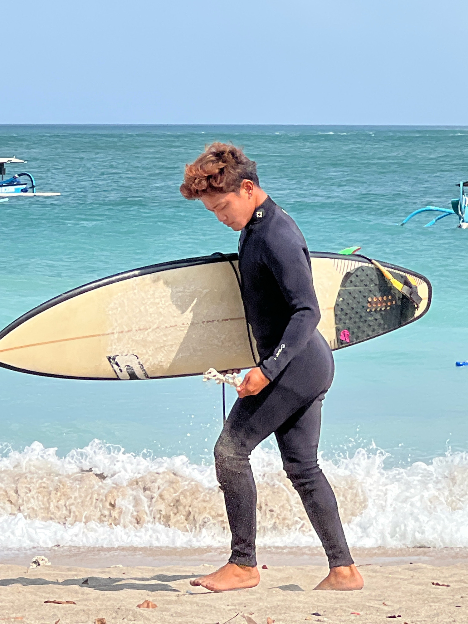 An Indonesian male surfer in a black wetsuit walks on the sandy beach of Kuta, Indonesia, carrying a cream-colored surfboard. The surfboard has a black fin and a black grip pad on the tail, with a leash secured to it. The ocean behind him is a vibrant turquoise with small waves breaking on the shore. Two traditional Balinese fishing boats, known as jukungs, are visible in the background on the water. The sky is clear and blue.