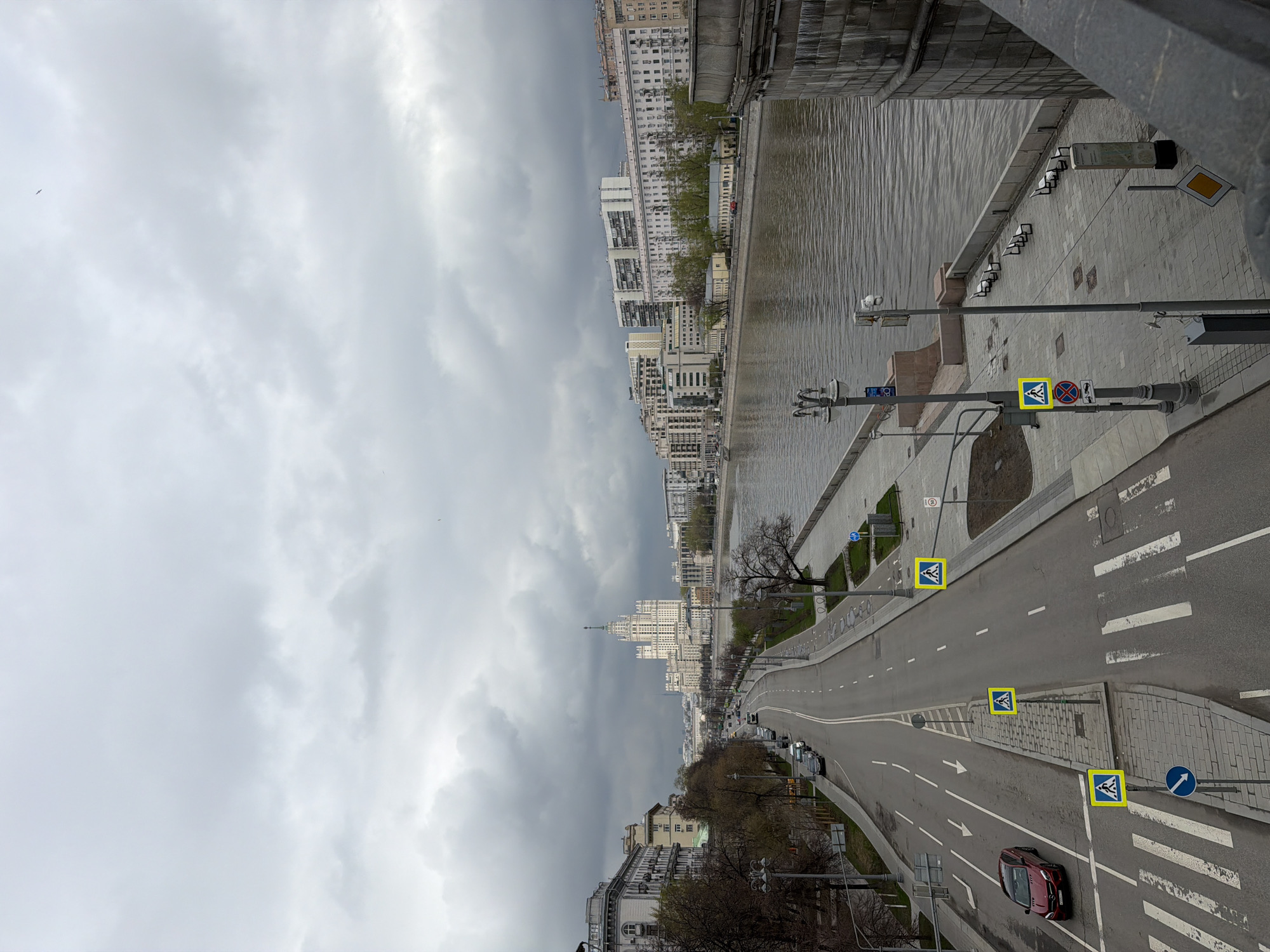 The image presents an elevated view of an urban landscape, likely Moscow, Russia, on an overcast day. The scene encompasses a wide river, a multi-lane road, and a skyline of city buildings under a dense, grey, cloudy sky.
In the foreground, a curving asphalt road dominates, featuring clearly marked lanes with white solid and dashed lines, turn arrows, and multiple pedestrian crosswalks. A dark red car is visible on the right side of the road. Several yellow diamond-shaped signs, indicating pedestrian crossings, are mounted on poles. Other visible road signs include a red circular "no stopping/parking" sign and a blue circular sign with a white arrow for mandatory direction. Streetlights line the road.
To the left of the road, a paved embankment borders a broad river, likely the Moskva River, whose surface is rippled and reflects the dull sky. The embankment includes a pedestrian walkway equipped with benches and lampposts. Across the river, a diverse array of buildings stretches into the distance. A prominent feature in the midground is a tall, ornate Stalinist-era skyscraper, identifiable as the Kotelnicheskaya Embankment Building, with its distinctive spire, towering above other residential and commercial structures along the riverbank. The trees along the road and river are mostly bare, suggesting late winter or early spring.
The overall impression is one of a vast, quiet urban environment, devoid of visible human activity, under a subdued, cloudy sky, highlighting the city's infrastructure and architecture.