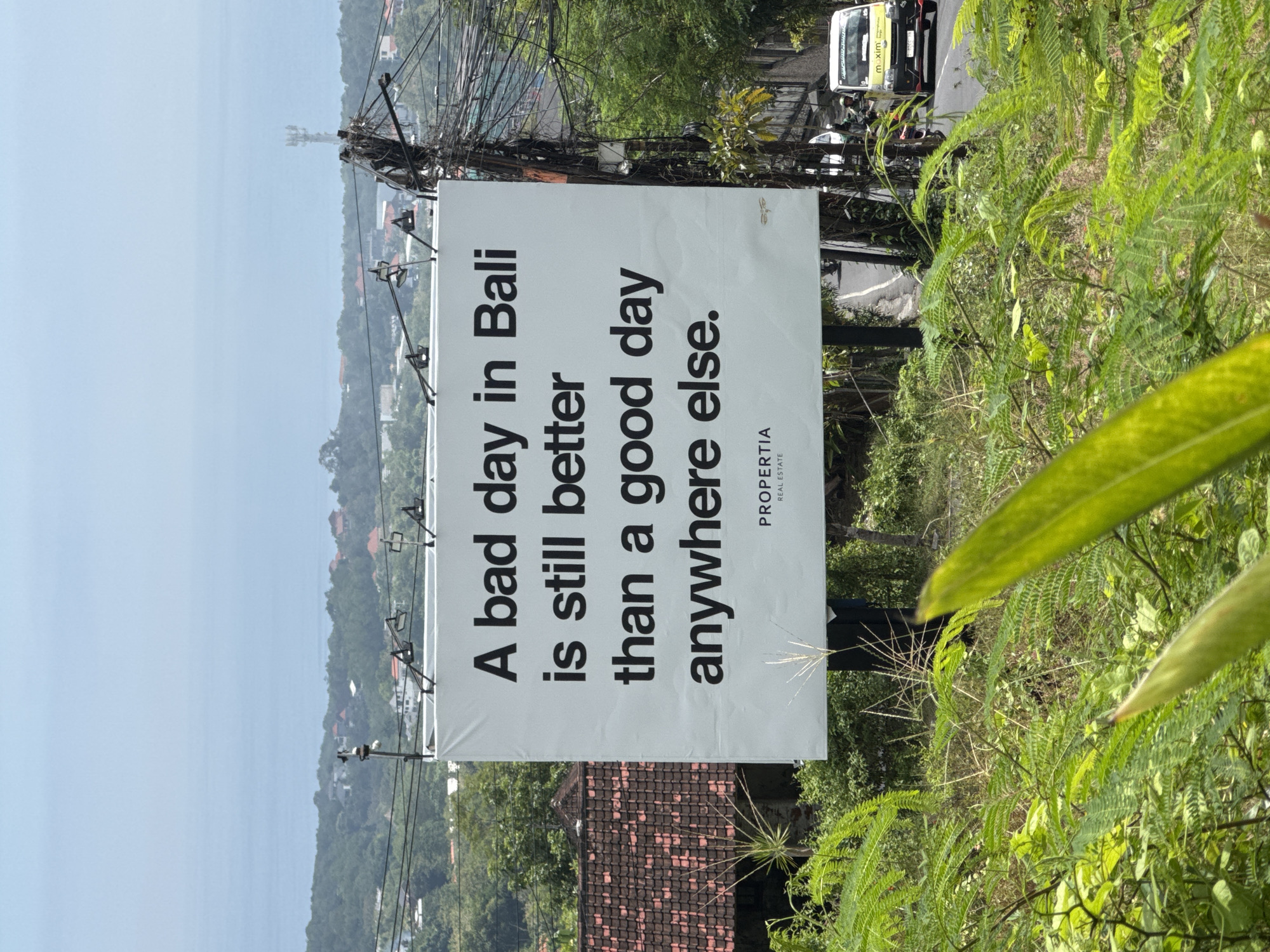 Here's a concise description of the image:
The image captures a billboard in Kuta Selatan, Indonesia, featuring the text "A bad day in Bali is still better than a good day anywhere else." The billboard promotes Propertia Real Estate. Overhead power lines crisscross above the billboard, and the background reveals a partial view of the ocean and surrounding landscape with a tiled-roof building and lush vegetation. The foliage appears to be growing nearby. A vehicle with a "maxim" logo is visible on the adjacent road. The photograph is taken from a lower angle, adding perspective.