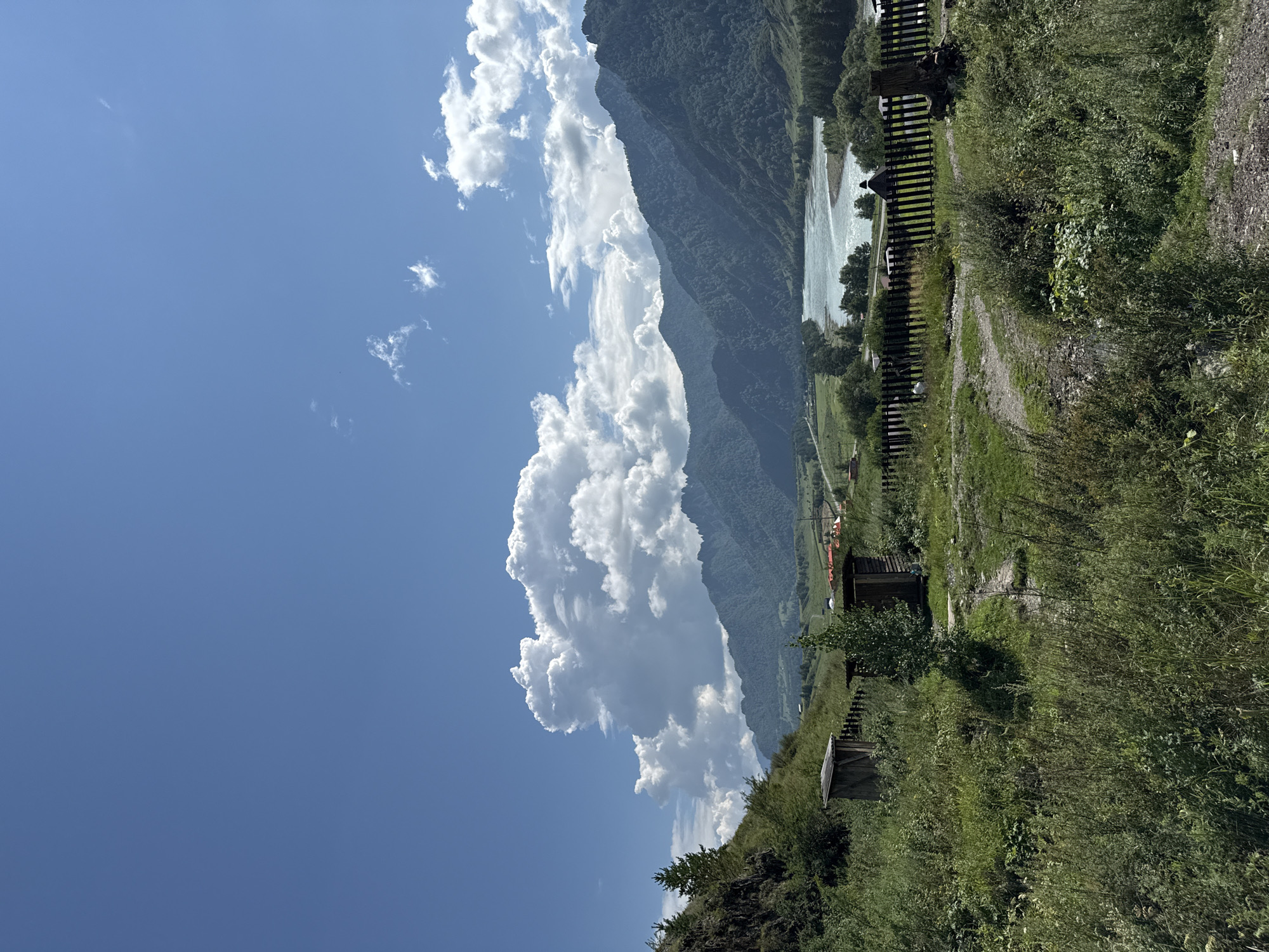 This is a landscape photograph taken in Chemal, Russia, featuring a bright blue sky with scattered cumulus clouds. In the foreground, a wooden fence runs horizontally across the scene, separating lush green vegetation and two small wooden outbuildings from the background. The outbuildings appear to be rustic and possibly primitive. Beyond the fence, a wide, turquoise river meanders through a valley, flanked by densely forested mountains that recede into the distance. The mountains are layered and have a rugged appearance. The overall impression is one of a natural, somewhat remote, and serene setting.