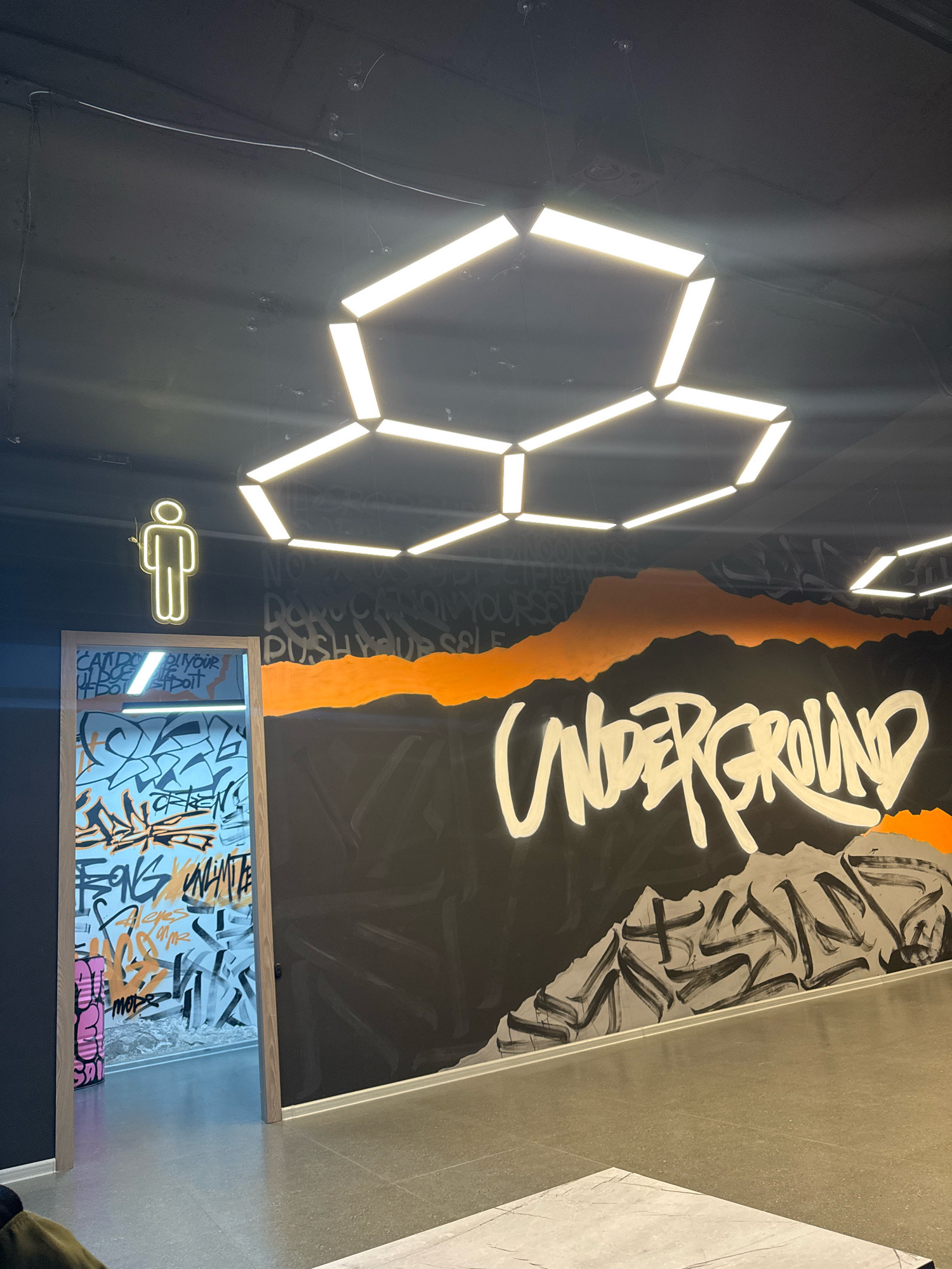 The image depicts an interior space with a dark ceiling featuring a geometric lighting fixture composed of hexagonal elements. A neon sign of a male figure is illuminated on the left wall. To the right of the neon sign, there is an open doorway leading into a room with walls covered in colorful graffiti. A large mural covers the wall to the right of the doorway, featuring the word "UNDERGROUND" in prominent white lettering against a black background with orange and grey abstract elements. The floor is a neutral grey tiled surface. This scene is located in the city of Almaty, Kazakhstan.