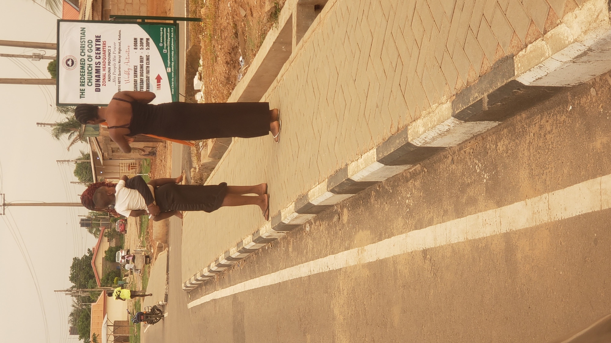 The image captures a daytime scene in Narayi, Nigeria, featuring "The Redeemed Christian Church of God, Dunamis Centre" sign. Two women are standing near the church's sign. One woman carries a child on her back, while the other appears to be an adult woman wearing a black dress. A road with a white line marking is visible in the foreground and cars, homes, and other structures of a typical city are shown in the background. There is a man on a bicycle in the road.