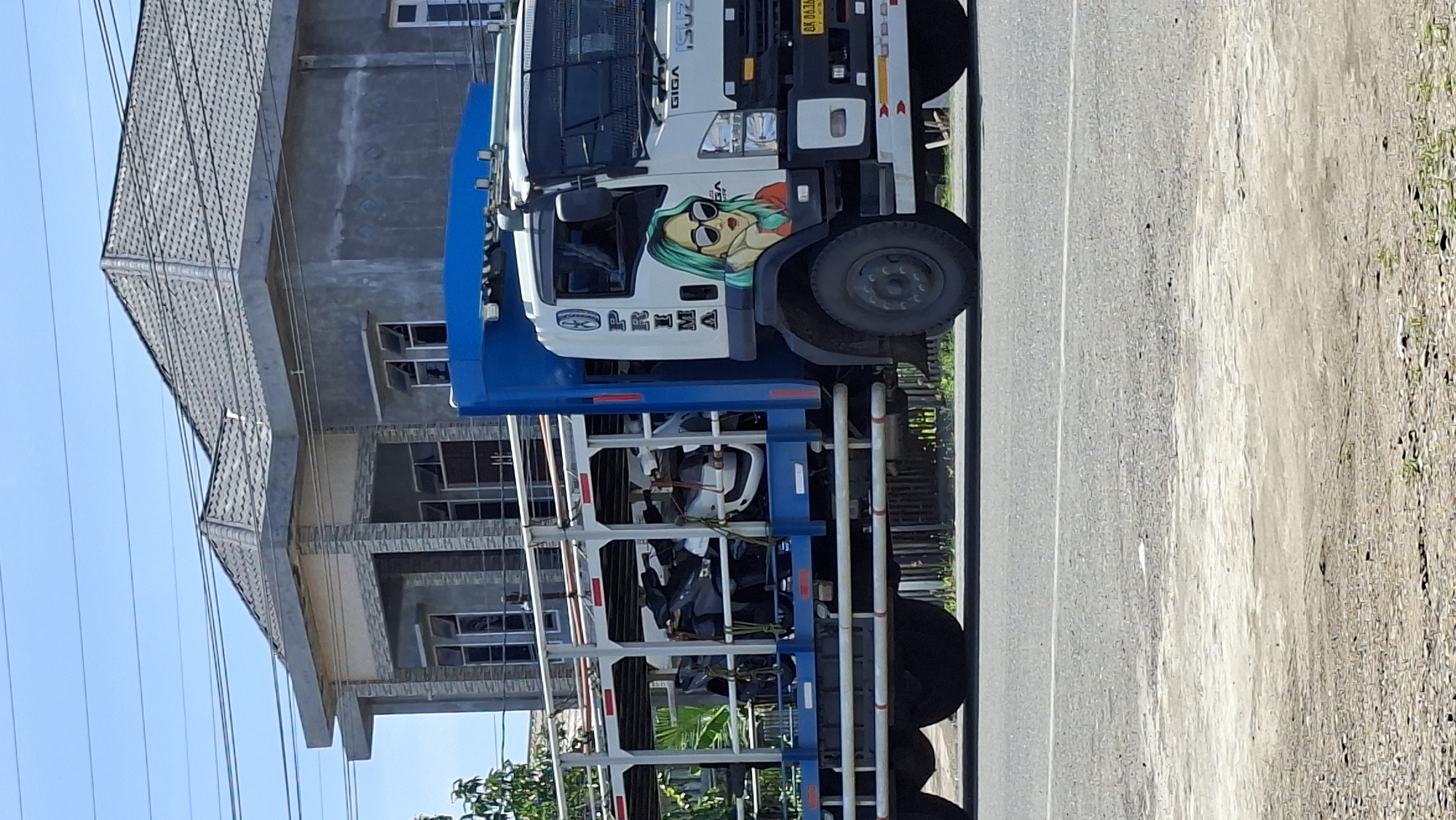 A side-view, eye-level shot captures a white and blue Isuzu truck transporting multiple motorcycles in Lhokseumawe, Indonesia, on a bright, clear day. The truck, with a "GIGA" logo visible, is decorated with a mural of a woman with green hair wearing sunglasses. The motorcycles are secured within a multi-level rack on the back of the truck. In the background, a modern, two-story house with a grey tiled roof and concrete walls is visible, suggesting a semi-urban or suburban setting. The truck is on a paved road, with power lines and vegetation also present in the scene.