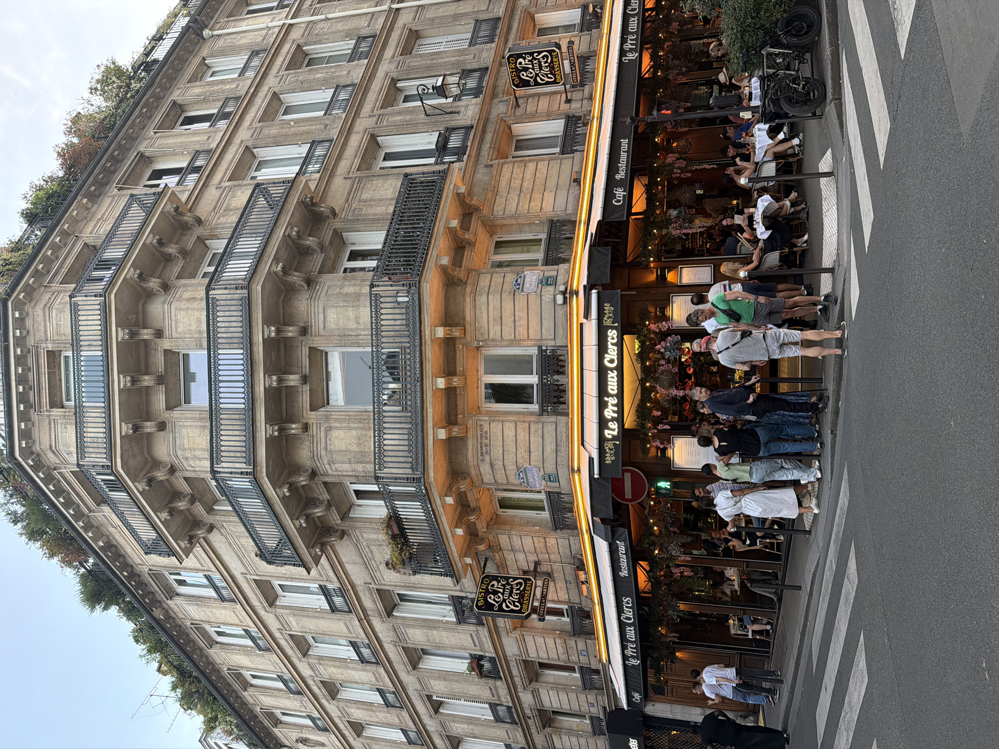 The image captures a bustling street scene in Paris, France, featuring the exterior of the "Le Pré aux Clercs" restaurant. The building has traditional Parisian architecture with stone facade, balconies, and window details. People are sitting at outdoor tables, enjoying meals and drinks under the restaurant's awning. Others are standing or walking along the sidewalk and crosswalk. The restaurant has a sign displaying its name and that it is a bistro, brasserie, cafe, and restaurant. The street appears busy with pedestrian traffic.
