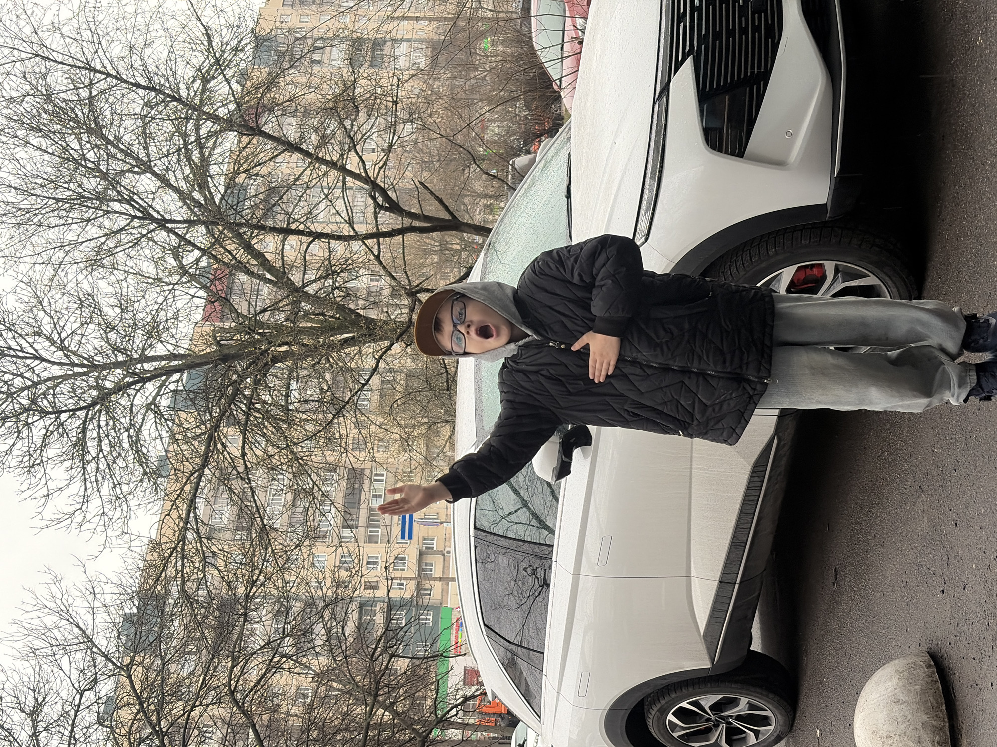 A young person is standing next to two white cars, with bare trees and a building in the background. The person is wearing a brown cap, glasses, a gray hooded sweatshirt, a black quilted jacket, and light gray pants. The person has their mouth open and is gesturing with both hands. The cars are parked on a wet paved surface. The building in the background has many windows and appears to be residential. The environment appears urban and the weather overcast. The location is not discernible as Saint Petersburg, Russia, from visual information in the image.