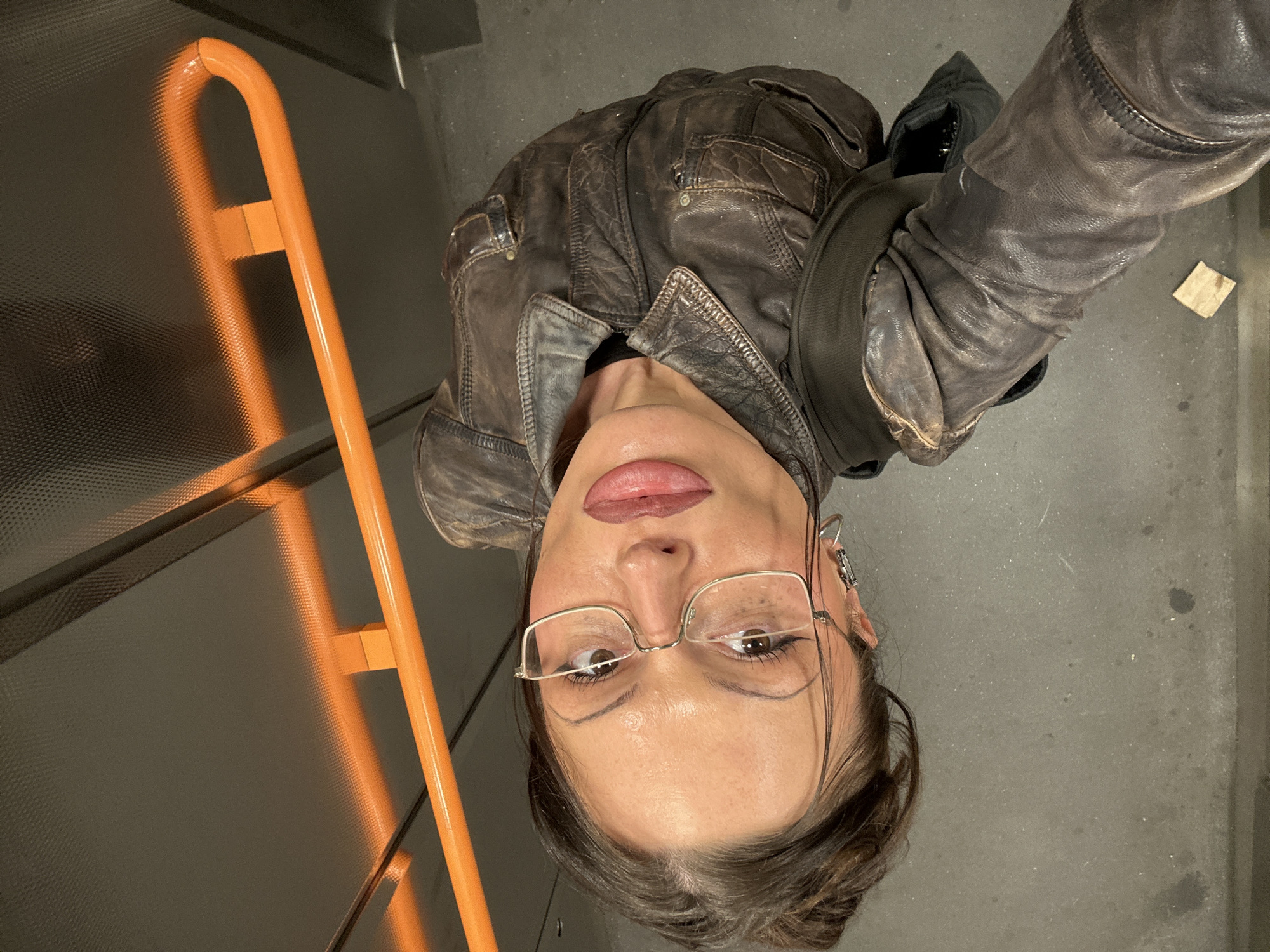 A medium shot, taken at an upward angle, shows a person in what appears to be a passenger elevator. The person is wearing glasses and a distressed brown leather jacket. An orange support bar for passengers is visible on the left, attached to the elevator wall, which is textured. The floor of the elevator is concrete. The location is implied to be in the city of London, United Kingdom, although there are no visible cues to confirm this definitively.