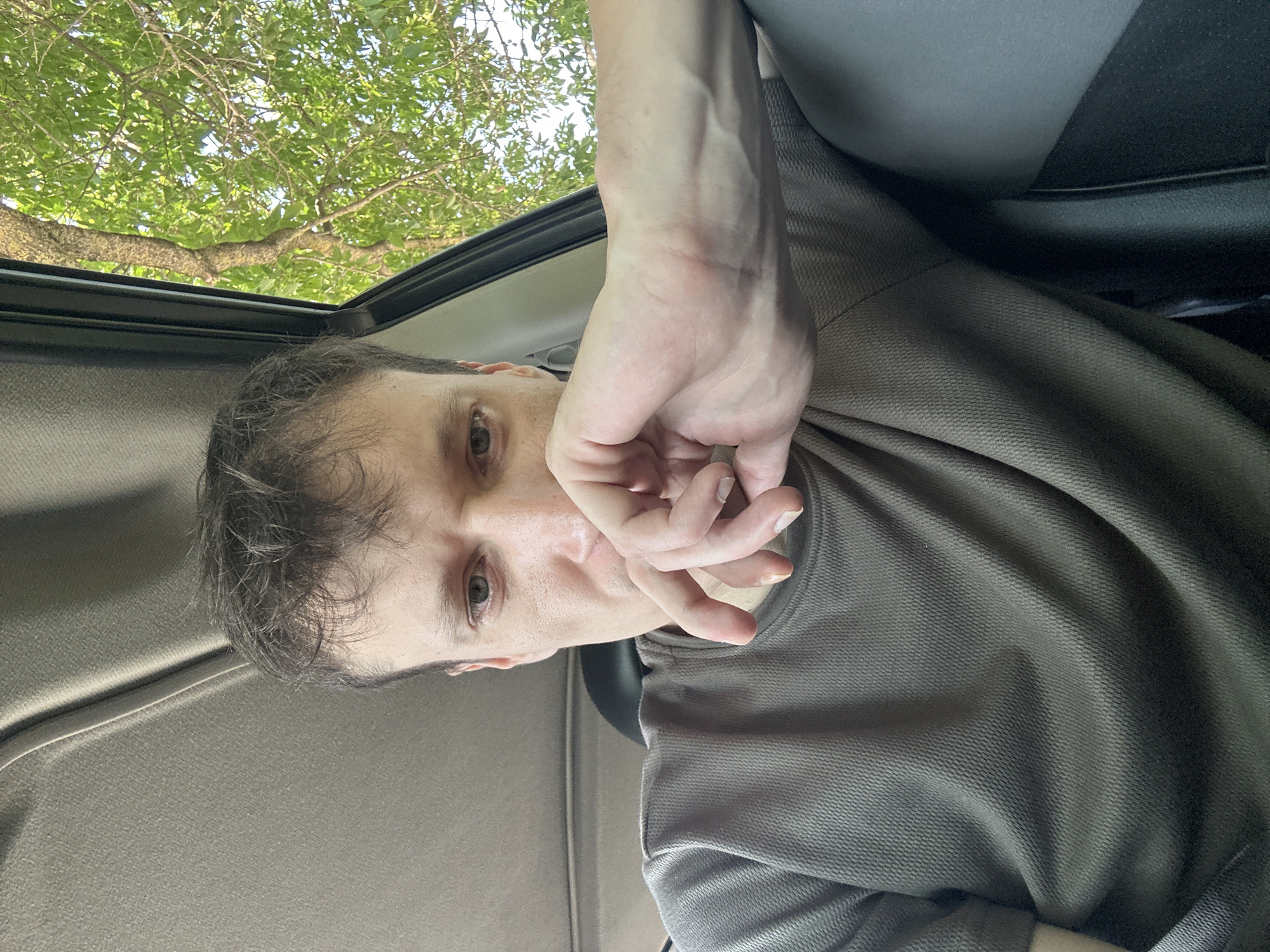 A man is sitting inside a car, resting his chin on his hand. He is wearing a dark gray t-shirt. The interior of the car, including the headliner and car seat, is visible. Outside the car window, a tree with green leaves can be seen. The image appears to have been taken during daylight hours. The location in the city of Makiv, Ukraine cannot be determined from the provided image.