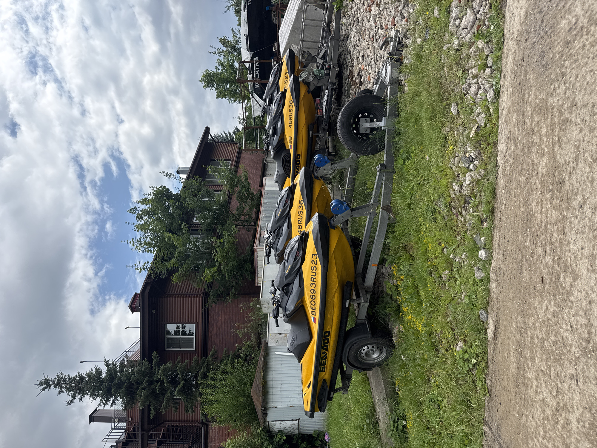 The image depicts a dry dock area in Povedniki, Russia, featuring a row of three yellow Sea-Doo personal watercraft parked on trailers. The watercraft are positioned side-by-side, facing towards the right of the frame. The frontmost watercraft displays the registration "BE0693RUS23" and the "SEA-DOO" logo. Behind it, another watercraft has "46RUS36" visible. The background consists of a two-story wooden building with a dark red facade, several trees, and a cloudy sky. The ground is covered with grass, gravel, and a concrete path in the foreground. There are no people visible in the image. The scene appears to be an outdoor storage or maintenance area for recreational vehicles.
