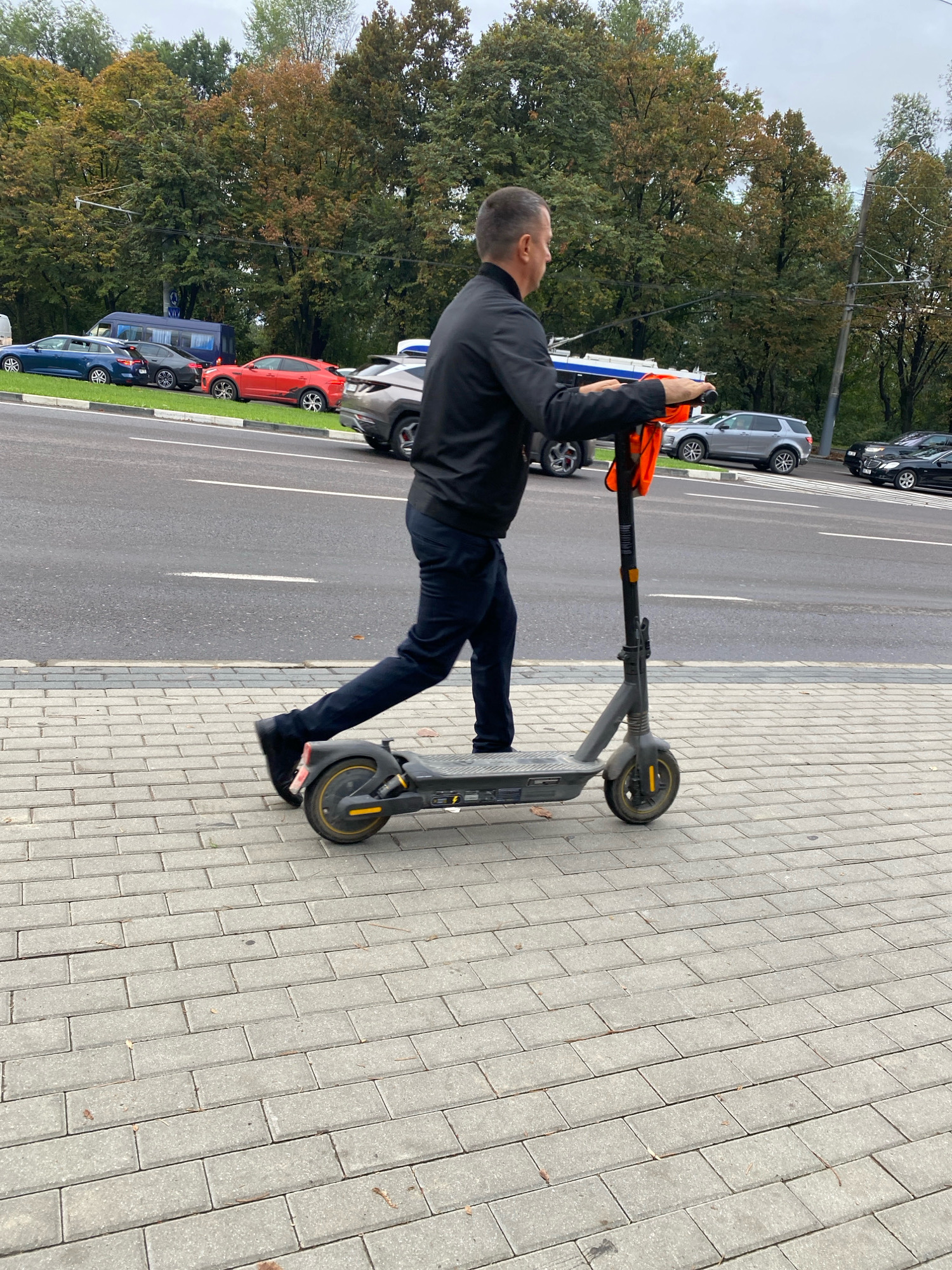 A man is shown from the side, riding a gray electric scooter on a paved sidewalk next to a road. He is wearing a black jacket and dark pants. An orange vest is attached to the scooter's handlebar. Several cars are parked and driving on the road behind him, and a line of trees with autumn foliage is visible in the background. The sky is overcast. The pavement is made of rectangular gray bricks. The scooter has two wheels, a platform, a handlebar, and a pole. The man's left leg is extended backward, pushing off the ground. The scene appears to be in an urban setting with street infrastructure.