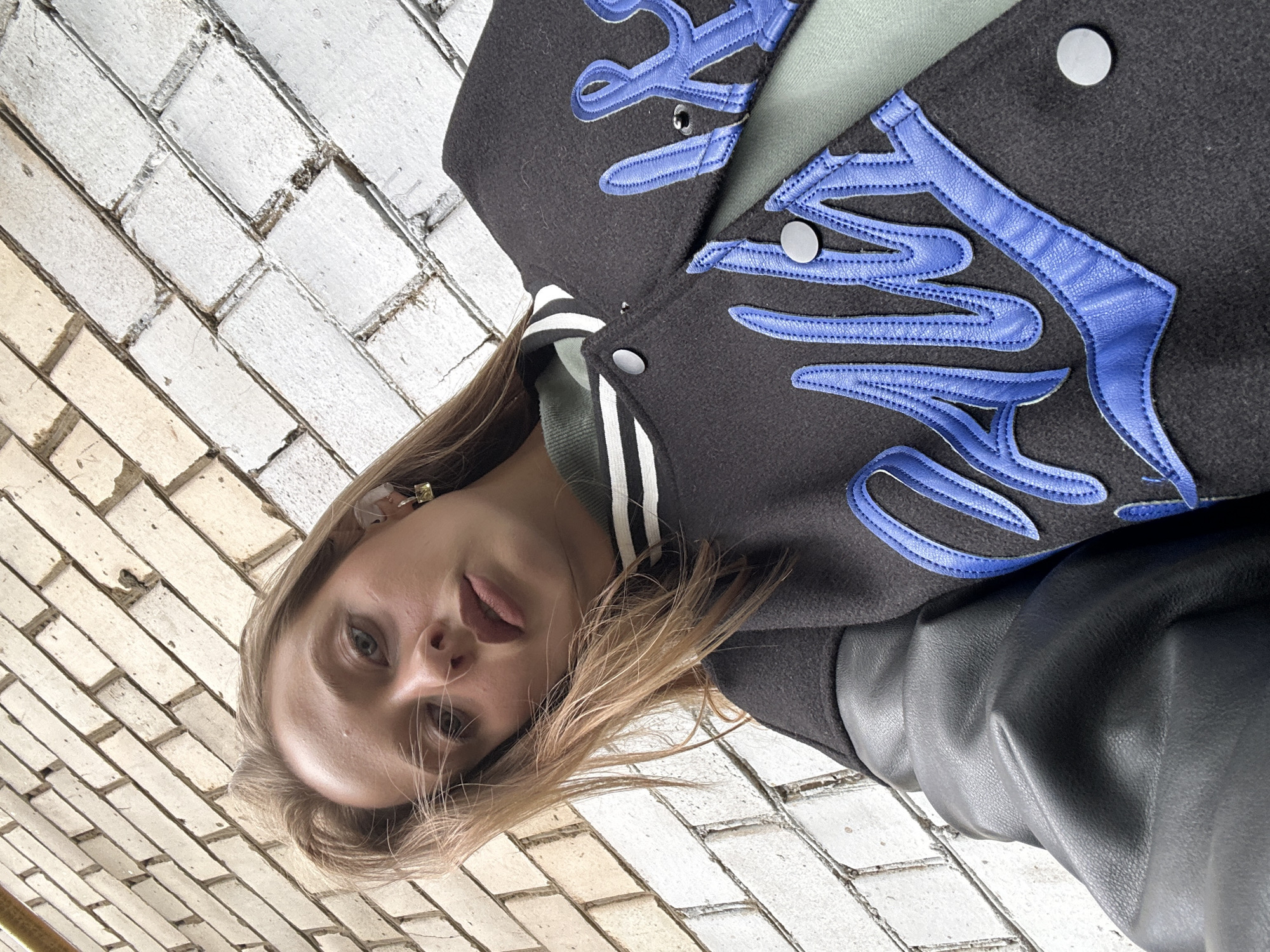 A young woman with blonde hair and light blue eyes is posing for a selfie against a brick wall. She is wearing a dark bomber jacket with blue lettering and a green shirt underneath. The brick wall is composed of light tan and white bricks. The image was taken in Khimki, Russia.