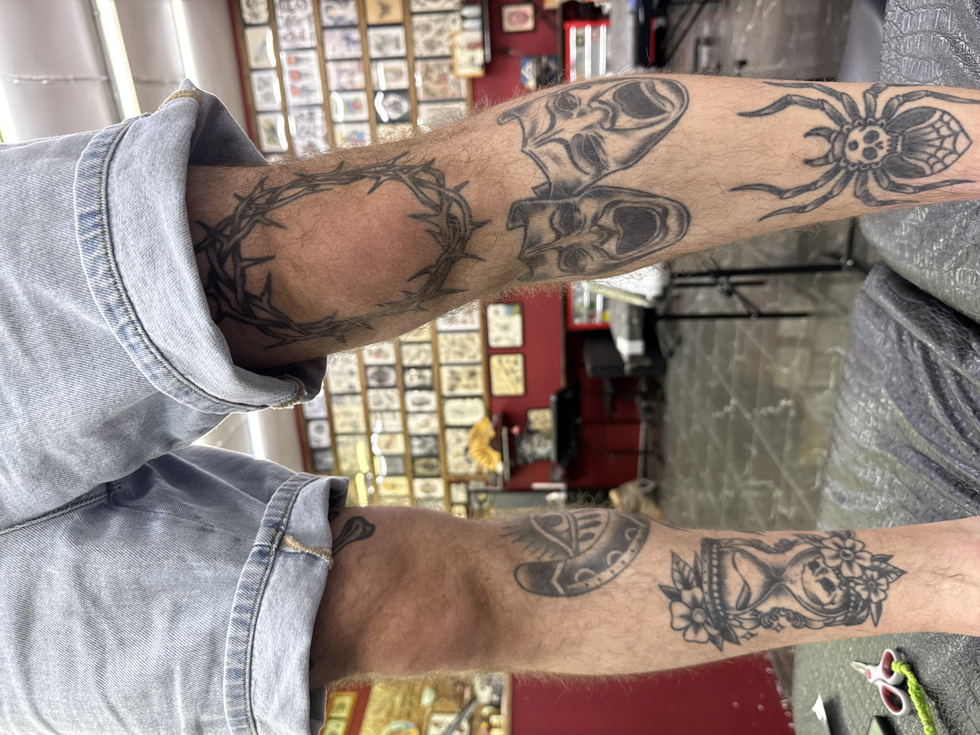 The image shows a close-up of a person's legs covered in tattoos, standing in what appears to be a tattoo parlor. The person is wearing denim shorts. The tattoos visible include a crown of thorns on the right thigh, theater masks on the right calf, an hourglass with a skull and flowers on the left calf, and a spider on the lower right leg. In the background, numerous framed tattoo designs are displayed on the walls. There is also a red counter and various pieces of equipment typical of a tattoo studio. The floor is tiled. The scene is a forensic, matter-of-fact depiction of the person's legs and tattoos within the environment of the tattoo parlor. There is no information provided within the image or context to indicate the location in Moscow, Russia.
