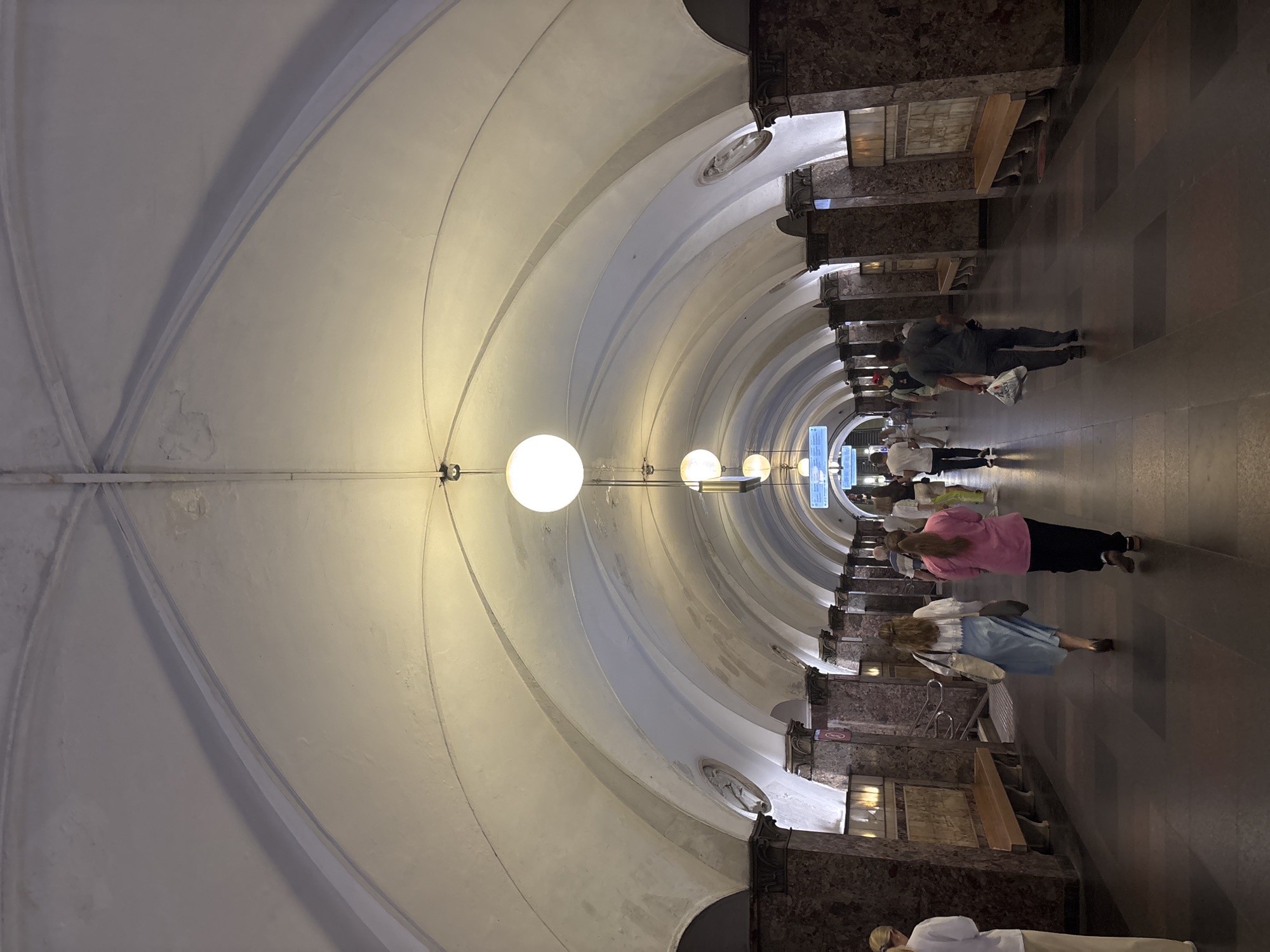 The image shows a long, arched corridor of what appears to be a metro station in Khimki, Russia. The ceiling is high and vaulted, with recessed lighting fixtures providing illumination. The walls are lined with marble or a similar stone, with decorative columns and benches along the sides. Several people are walking in both directions down the corridor, some carrying bags. The floor is tiled in a dark color with some lighter square accents. Signs are visible in the distance, indicating direction or information. The overall atmosphere is one of a busy public transit space.