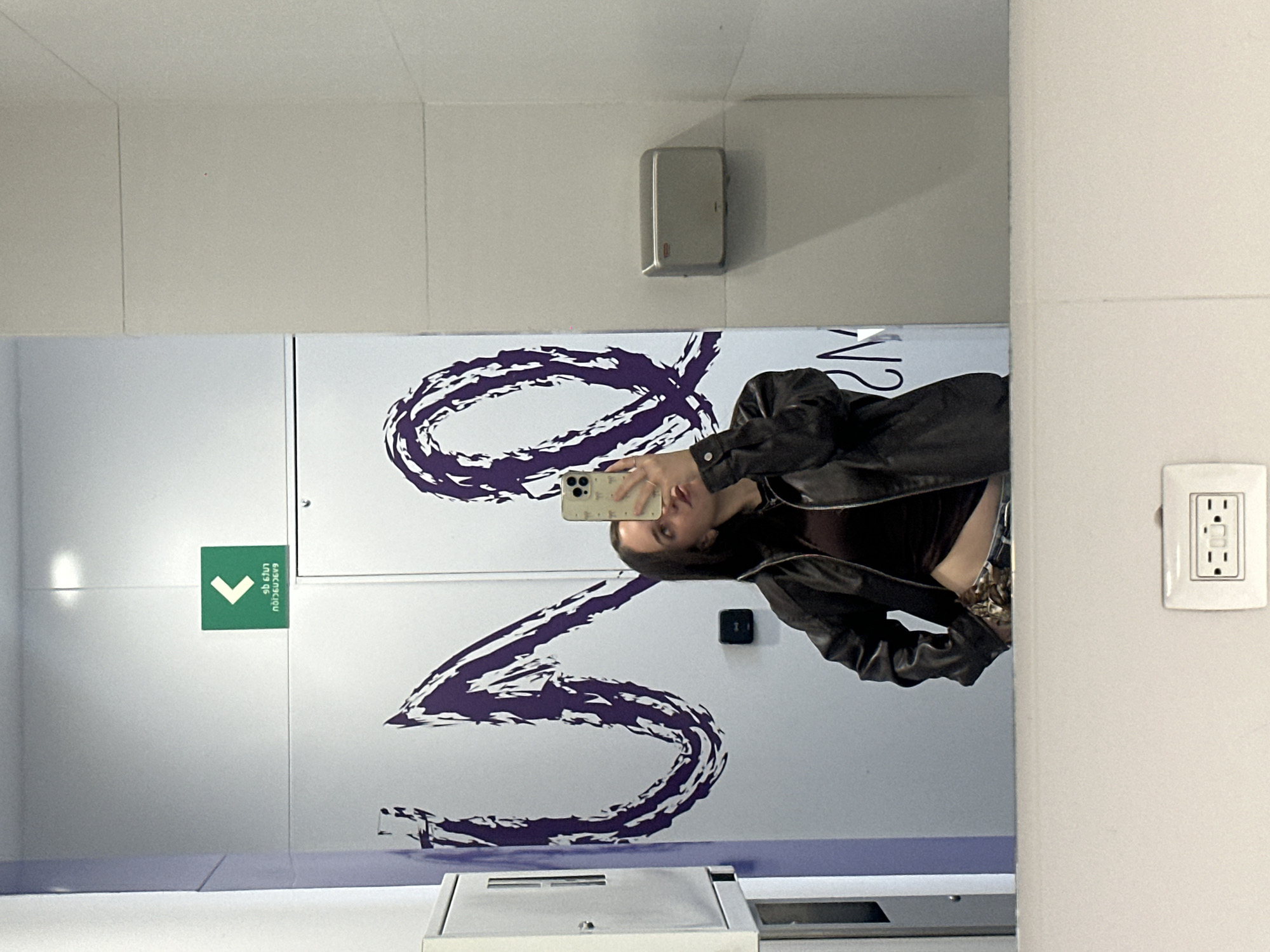 The image shows a mirror selfie in a restroom. A woman is holding a smartphone, taking the picture. The restroom features a white wall with a large, stylized purple graphic resembling a ribbon or a symbol. There is a green sign with a downward arrow and text in Thai. An outlet is visible on the wall. The location is Mexico City, Mexico.
